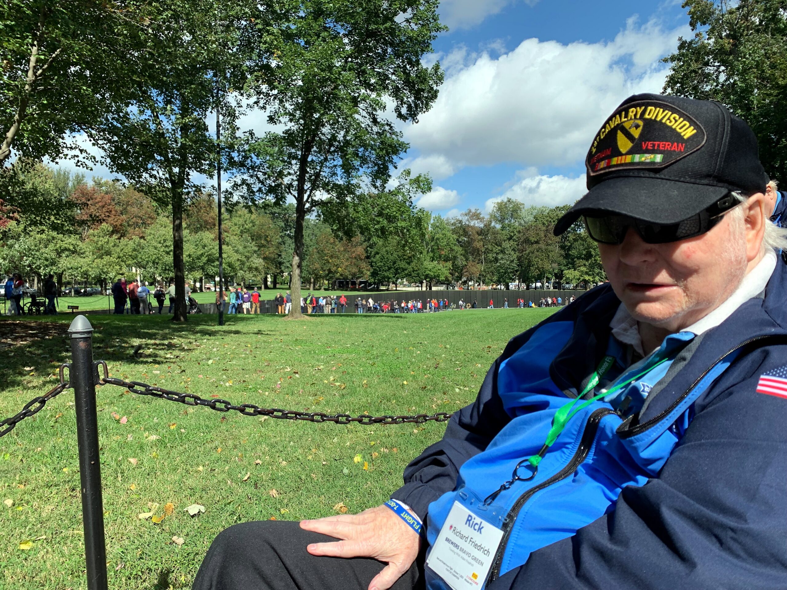 Close-up of an older man wearing a black Veteran hat and sunglasses, sitting in a park with a chain fence and a crowd in the distance on a sunny day.