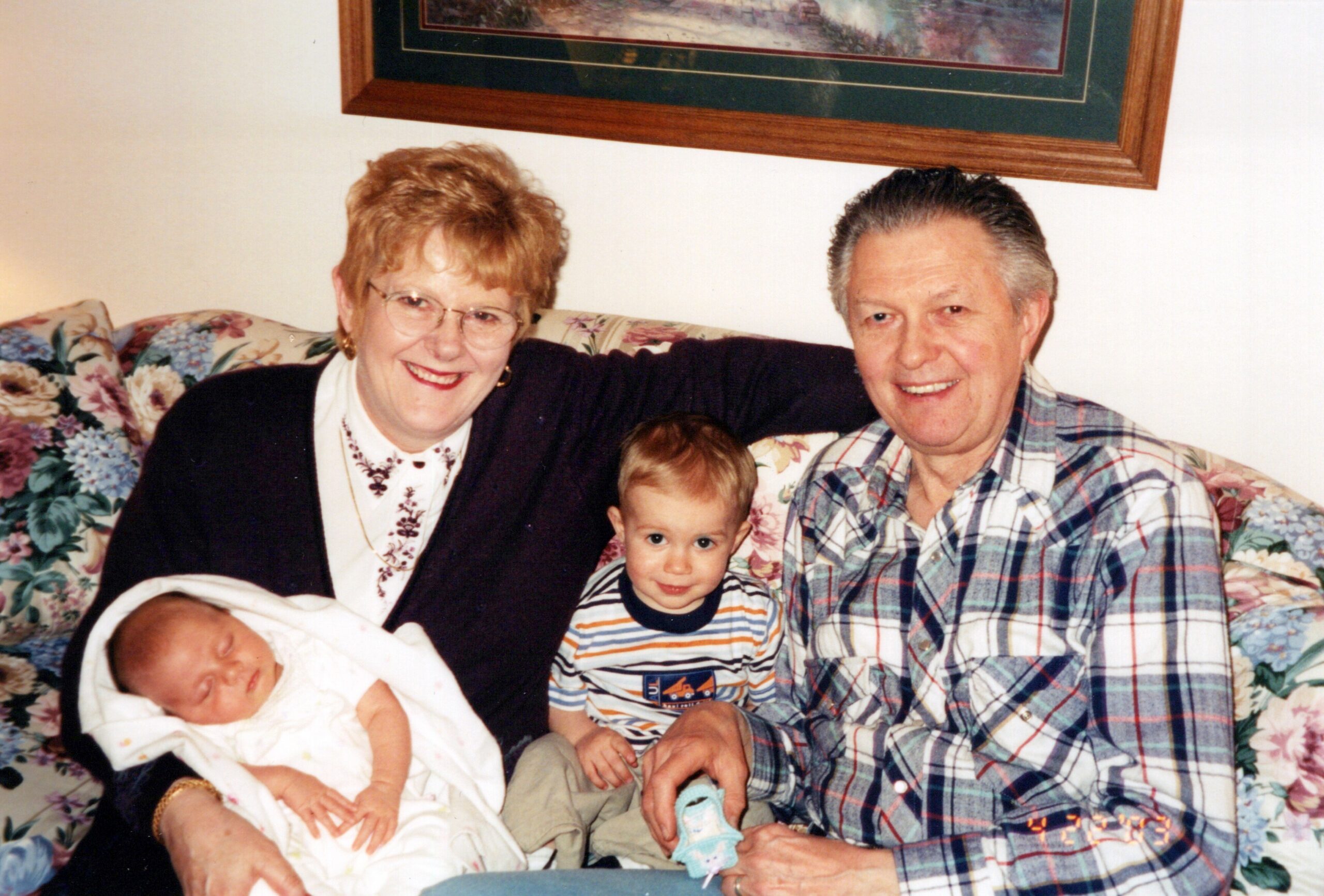 Smiling elderly couple on a floral couch with a baby wrapped in white and a young toddler between them.