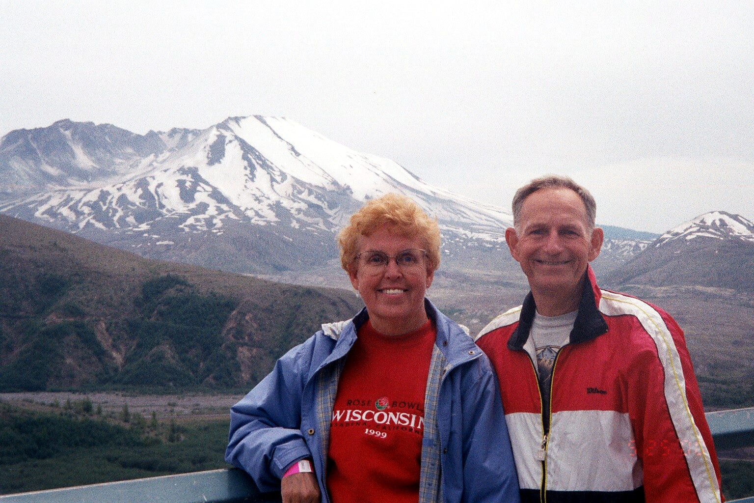Smiling couple in jackets pose at a railing with snow-capped mountains behind them, a scenic overlook.