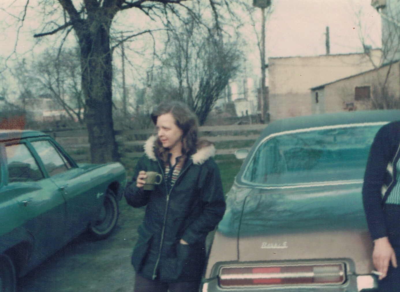 Person wearing a dark coat with a fur-lined hood holds a mug while standing between two vintage cars in a driveway.