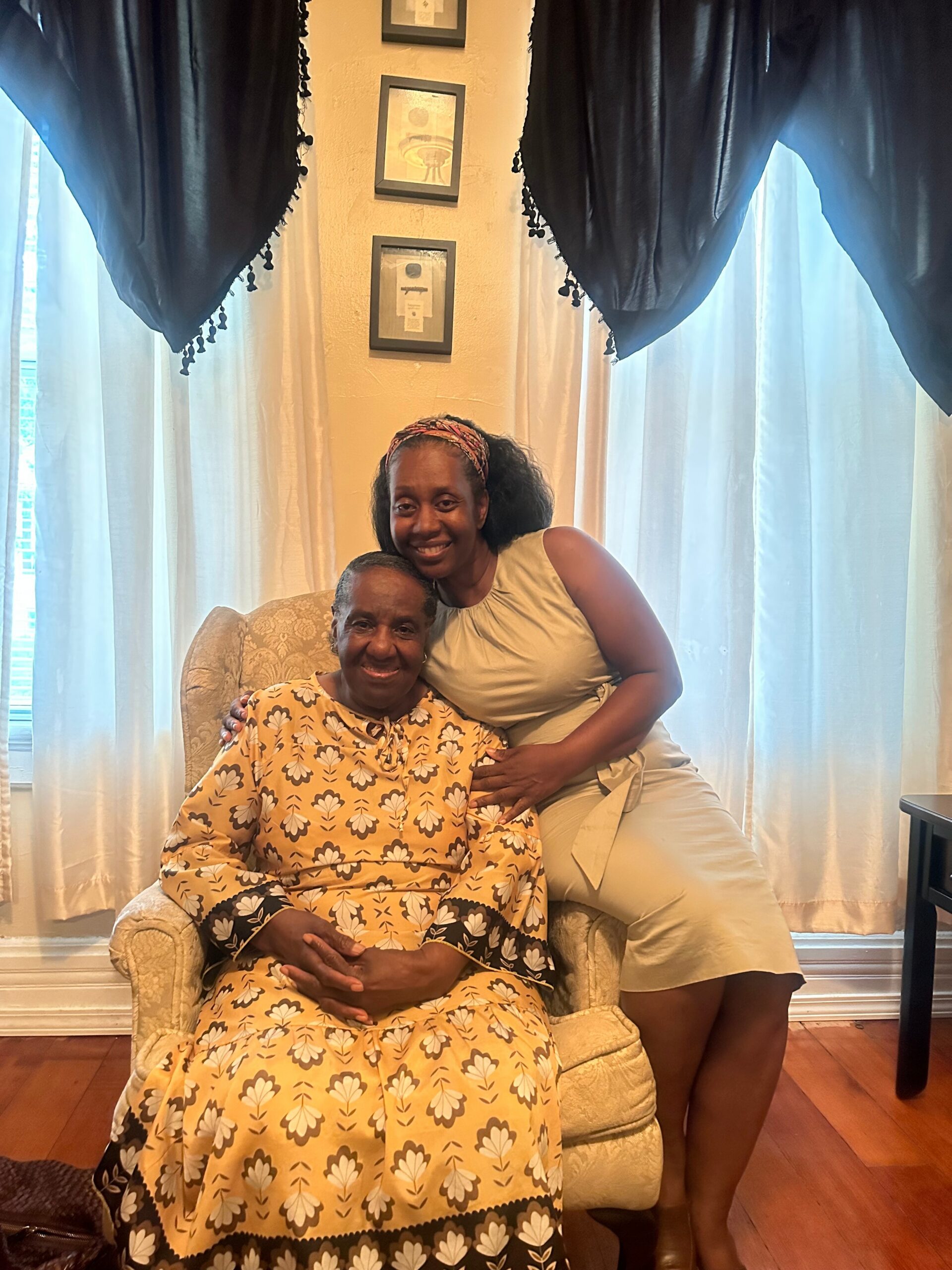 Older woman in a yellow floral dress sits in a patterned chair, smiling, as a younger woman stands beside her with an arm around her in a living room.