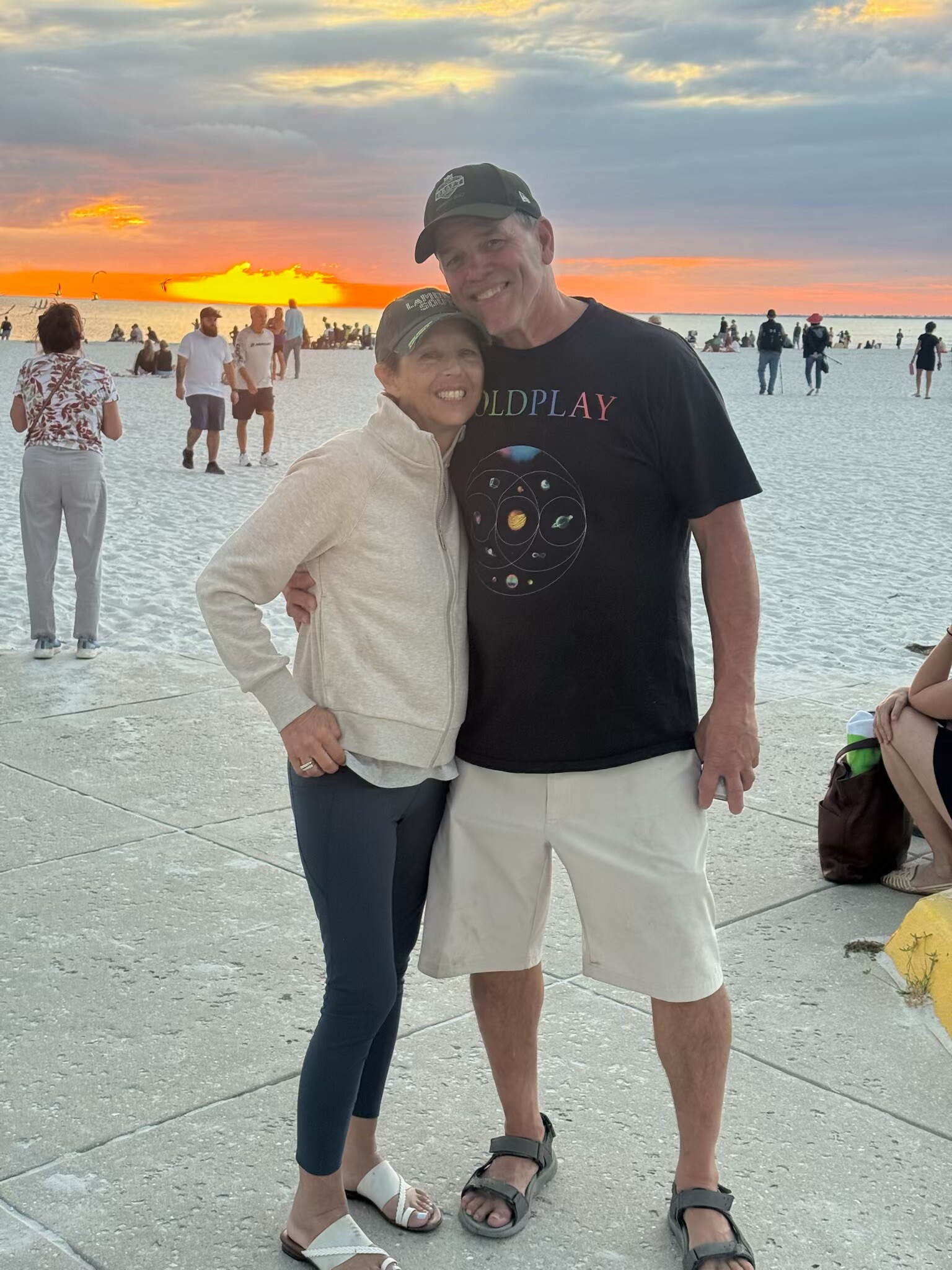 Smiling couple on a white-sand beach at sunset, arms around each other, with people in the background.