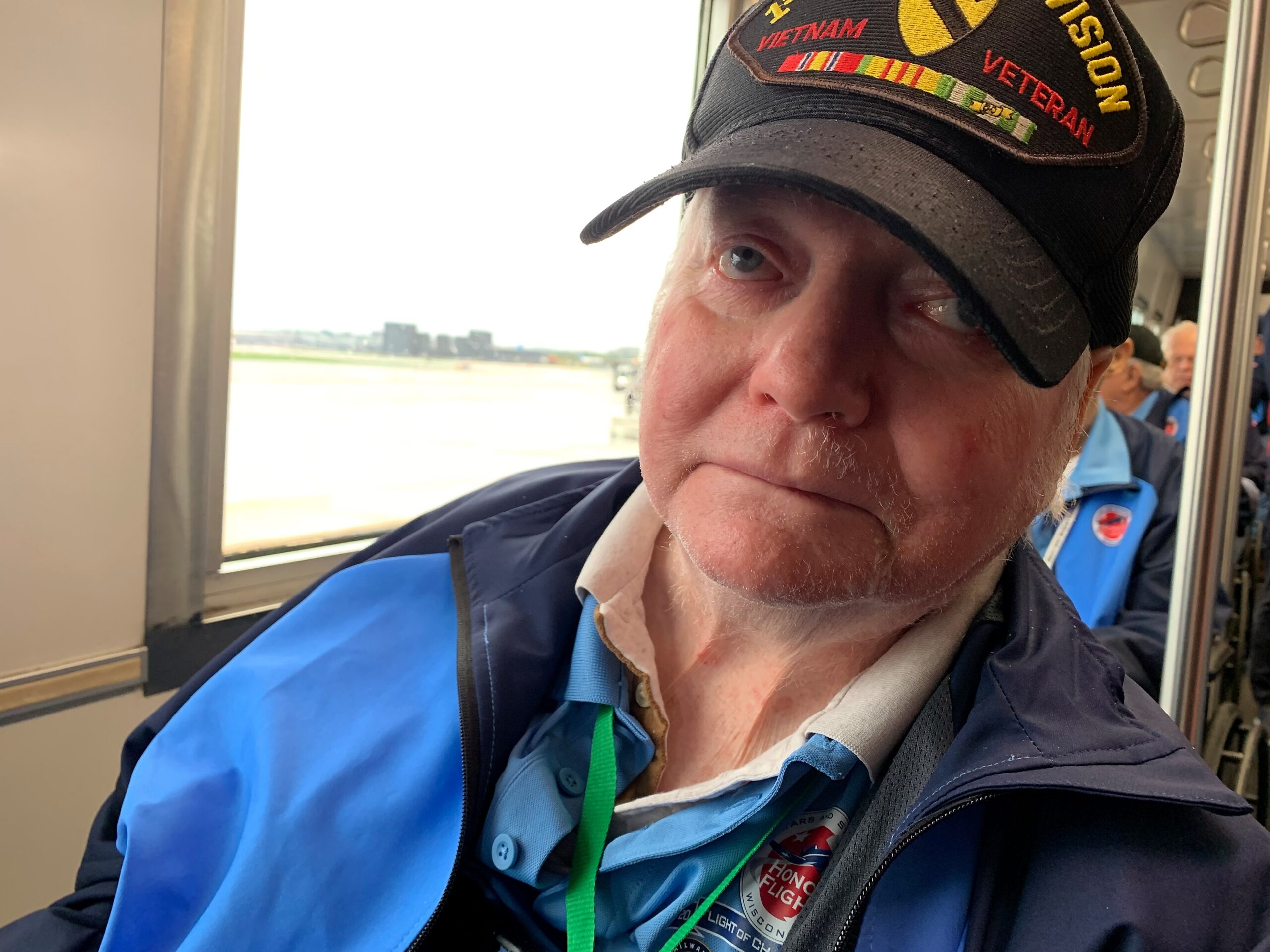 Close-up of a man wearing a black cap with a Vietnam Veteran patch, sitting on an airplane and looking aside.