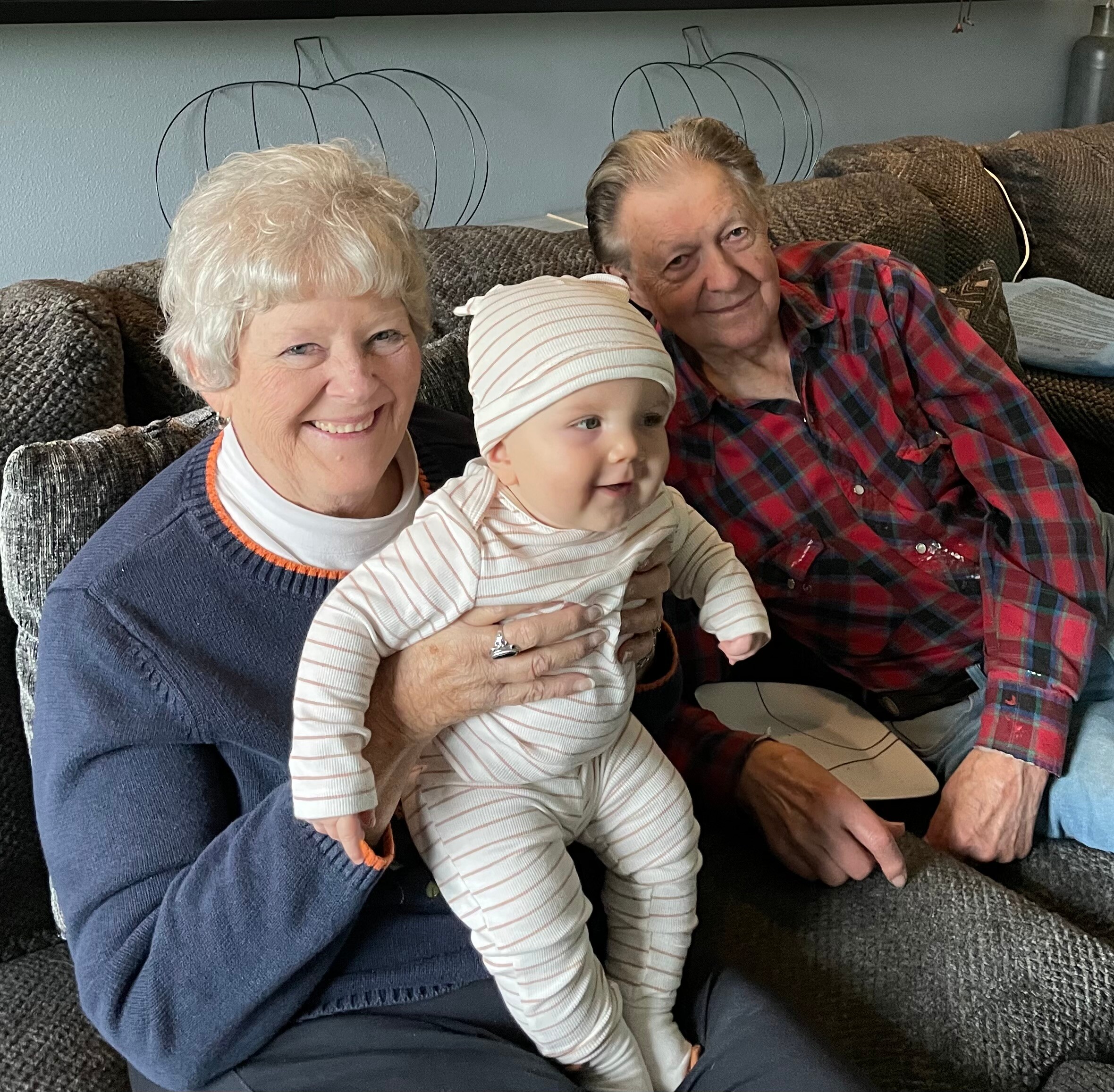 Three generations on a sofa: an elderly woman holding a smiling baby, with an older man in a red plaid shirt beside them.