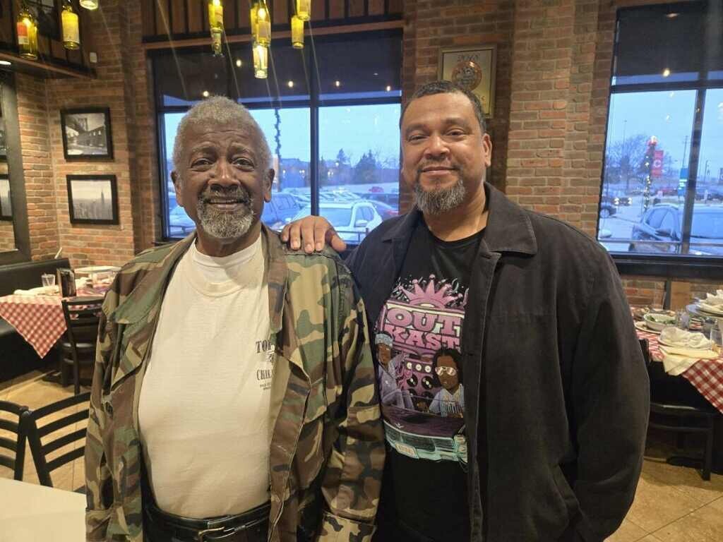 Two smiling men pose together in a brick-walled restaurant; one wears a camo jacket, the other a black jacket.