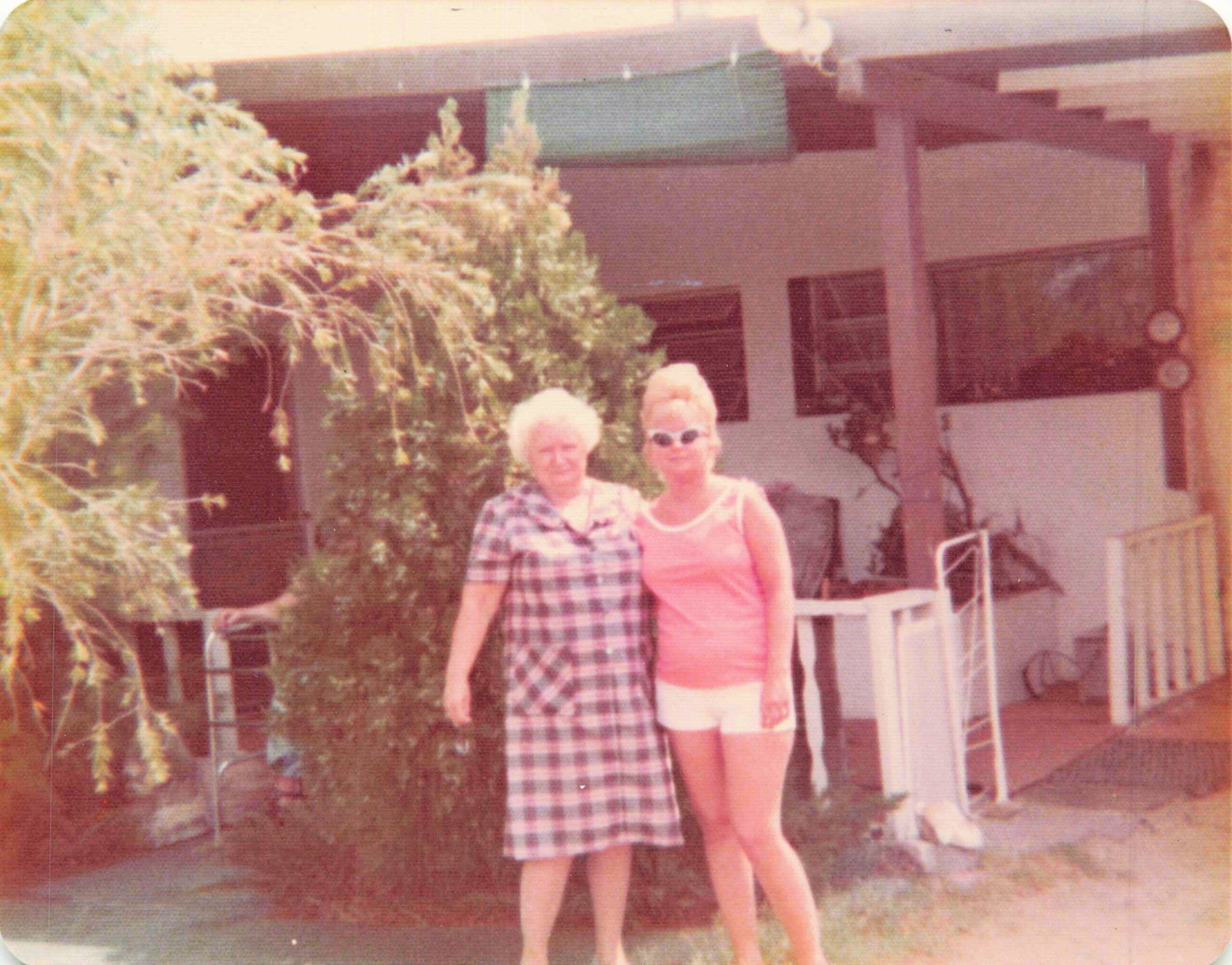 Two elderly women stand together in a yard in front of a house with a porch and steps, posing for a photo.