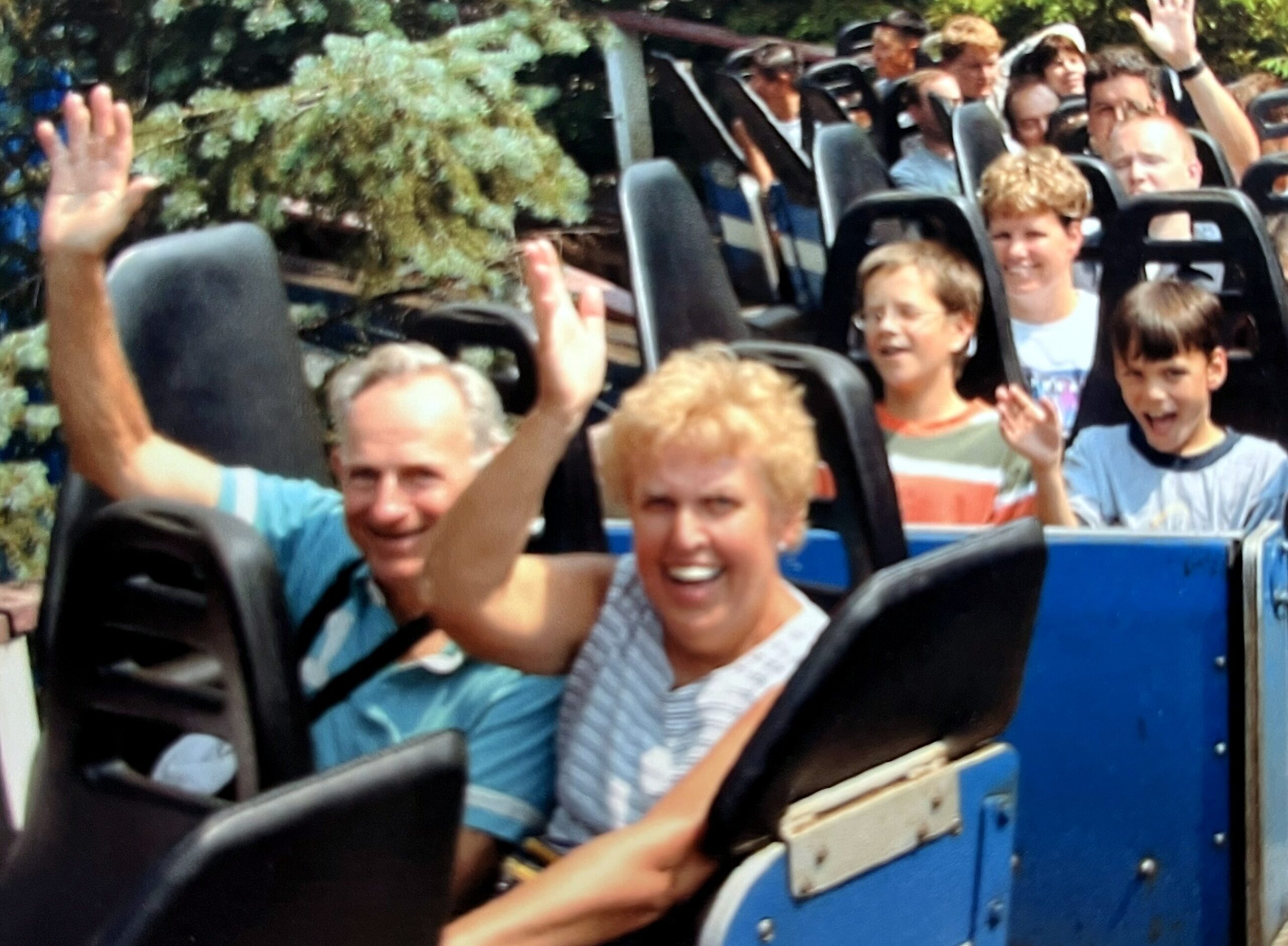 People on a roller coaster train with hands raised and smiles, blue track side panels visible in the foreground.