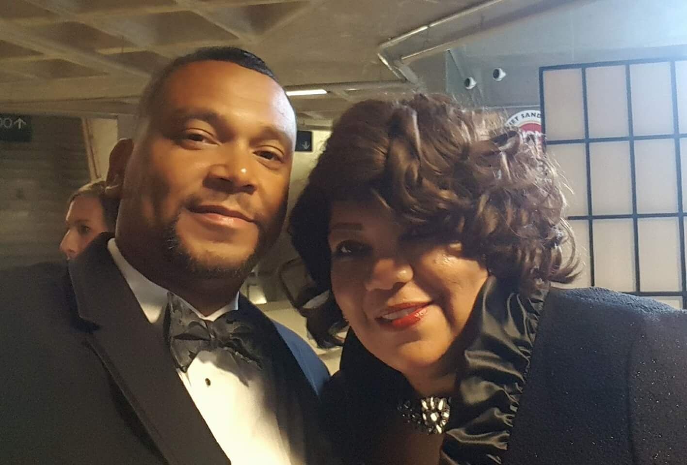 Two people in formal attire pose for a selfie in a backstage area, smiling at the camera.