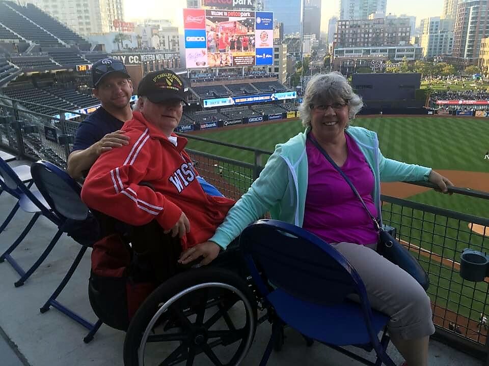 Three friends at a baseball stadium, two in wheelchairs, smiling with the field in the background. Two accessibility-friendly seats and a cheering crowd backdrop convey a joyful game-day moment.