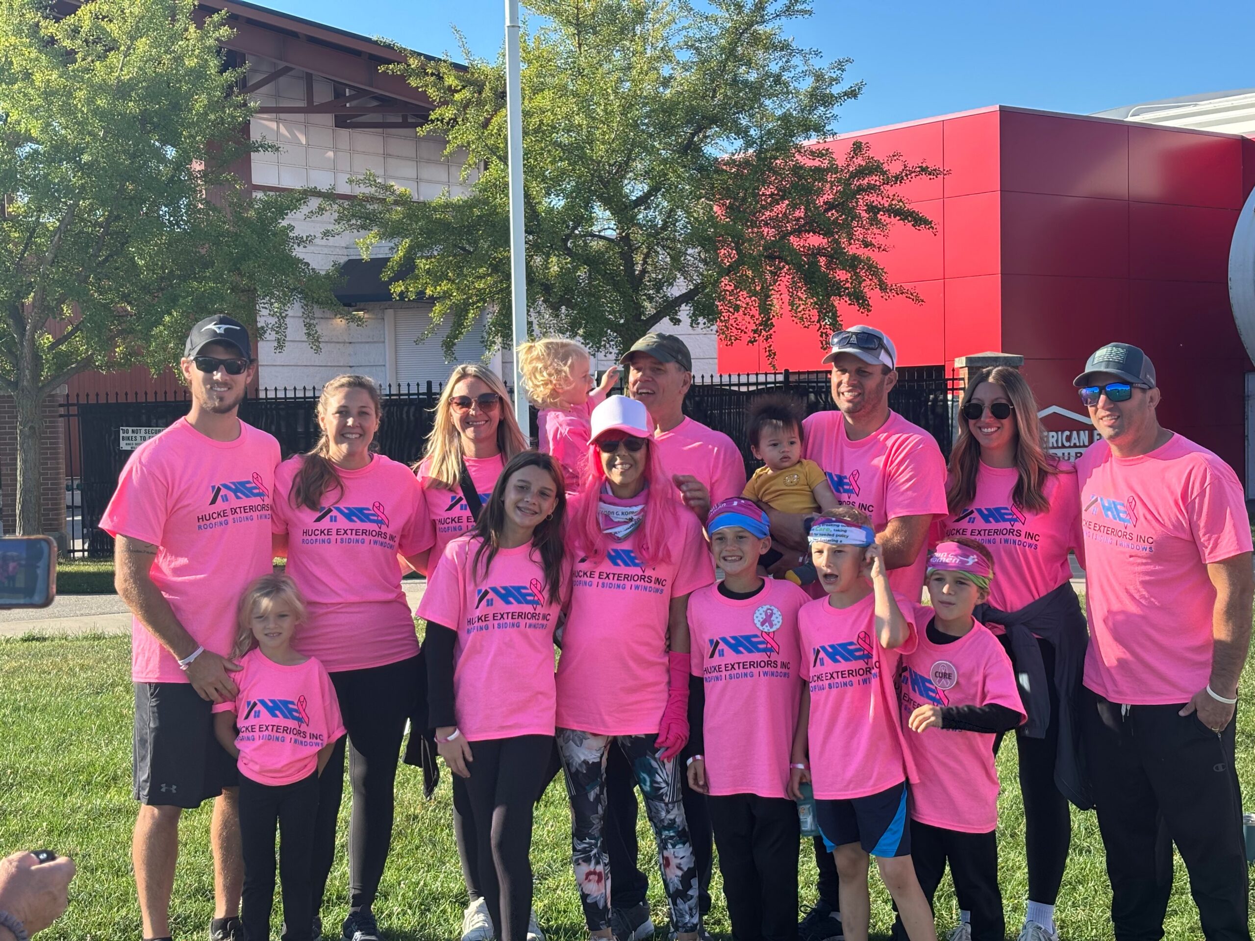 Group of people wearing bright pink shirts posing together outdoors, smiles for a team photo under a clear blue sky.
