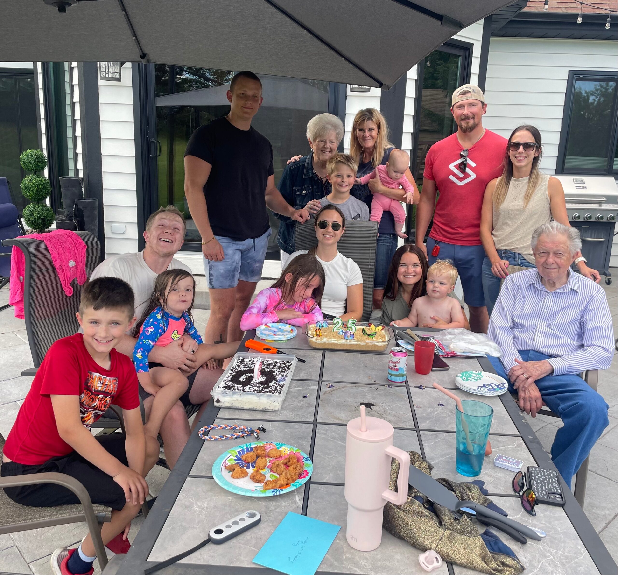 Family and friends gathered around a patio table for a birthday celebration outside, with cake and partyware visible.