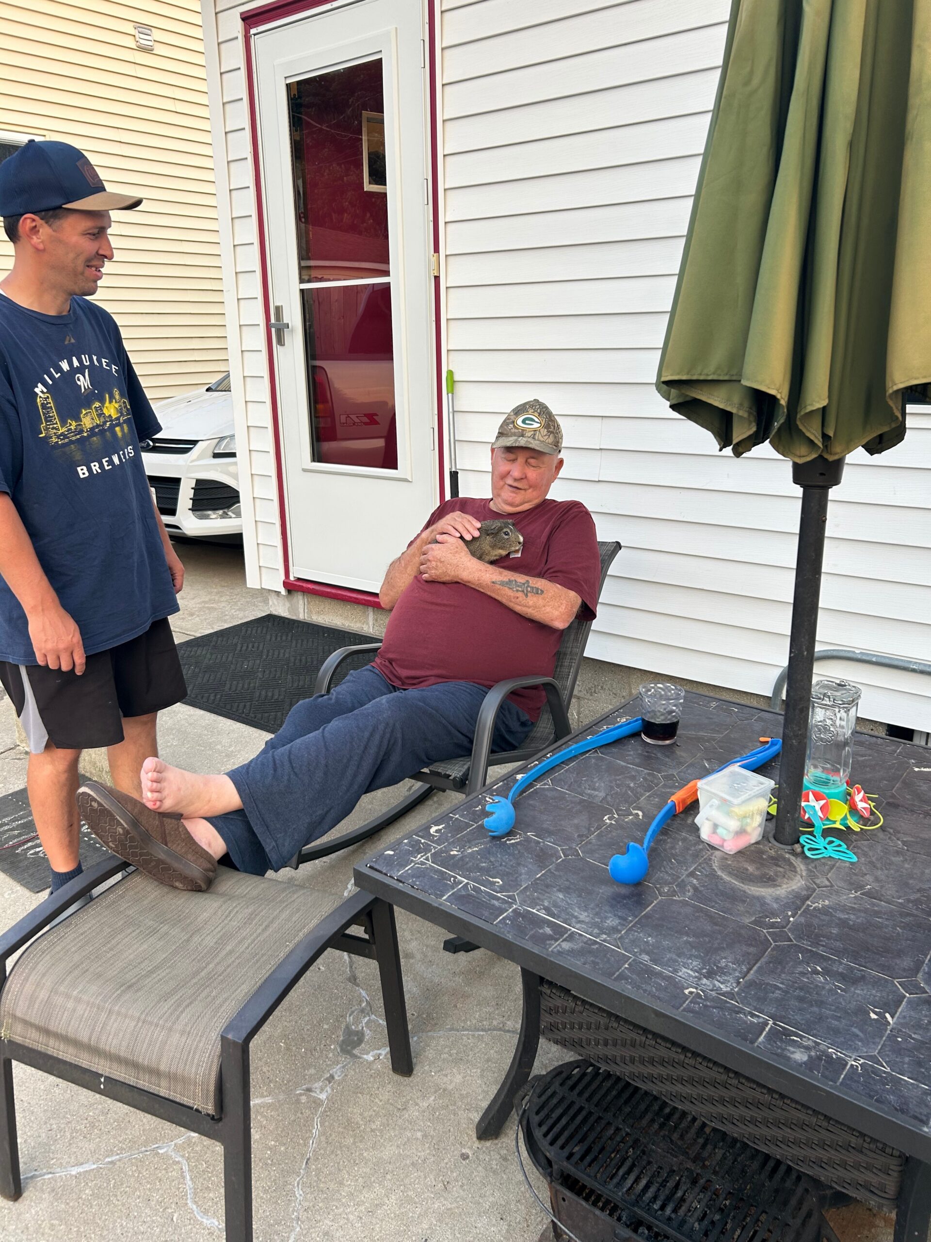 Two men outside a house; a man in a maroon shirt sits in a chair cradling a small gray rabbit while another man in a Milwaukee Brewers shirt stands nearby.