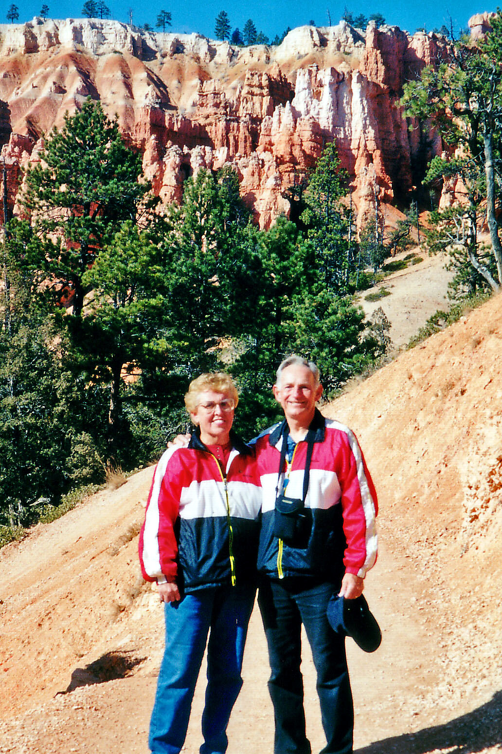 Older couple in red, white, and blue jackets posing on a dirt hiking trail with orange canyon cliffs and pine trees behind them.
