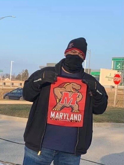 Person outdoors wearing a Maryland Terrapins shirt with the terrapin mascot, a black jacket, and a face covering to display the logo.