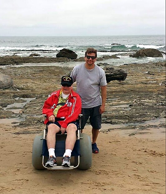 Man in a wheelchair on the beach being pushed by a friend, with rocky shoreline and waves in the background.