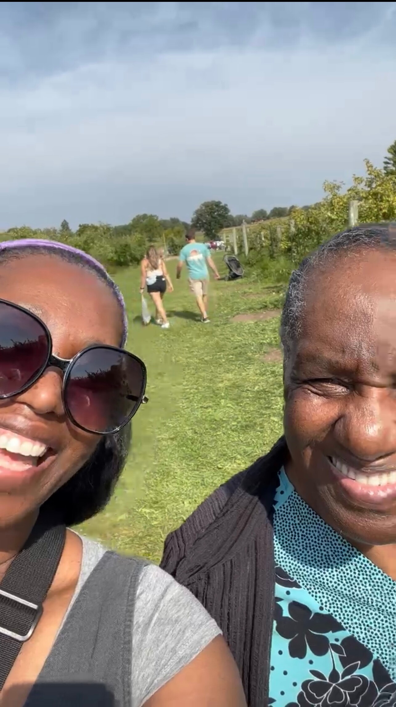 Two people in the foreground smiling for a selfie on a sunny day, with a grassy path and other walkers in the background.
