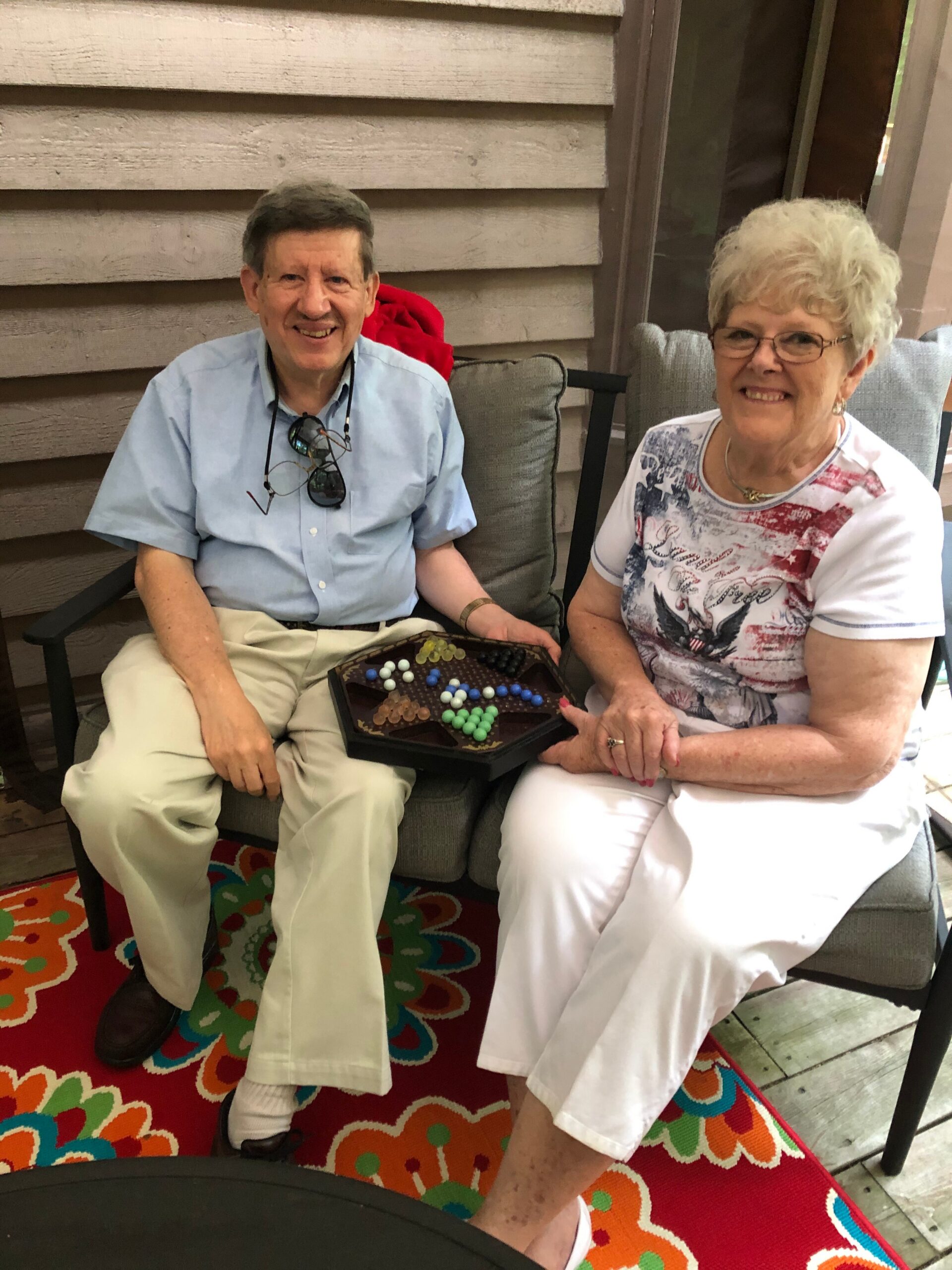 Two smiling seniors sit on a covered porch, a tray of colorful chocolates resting on the man's lap while he wears a light blue shirt and sunglasses hanging from his shirt.