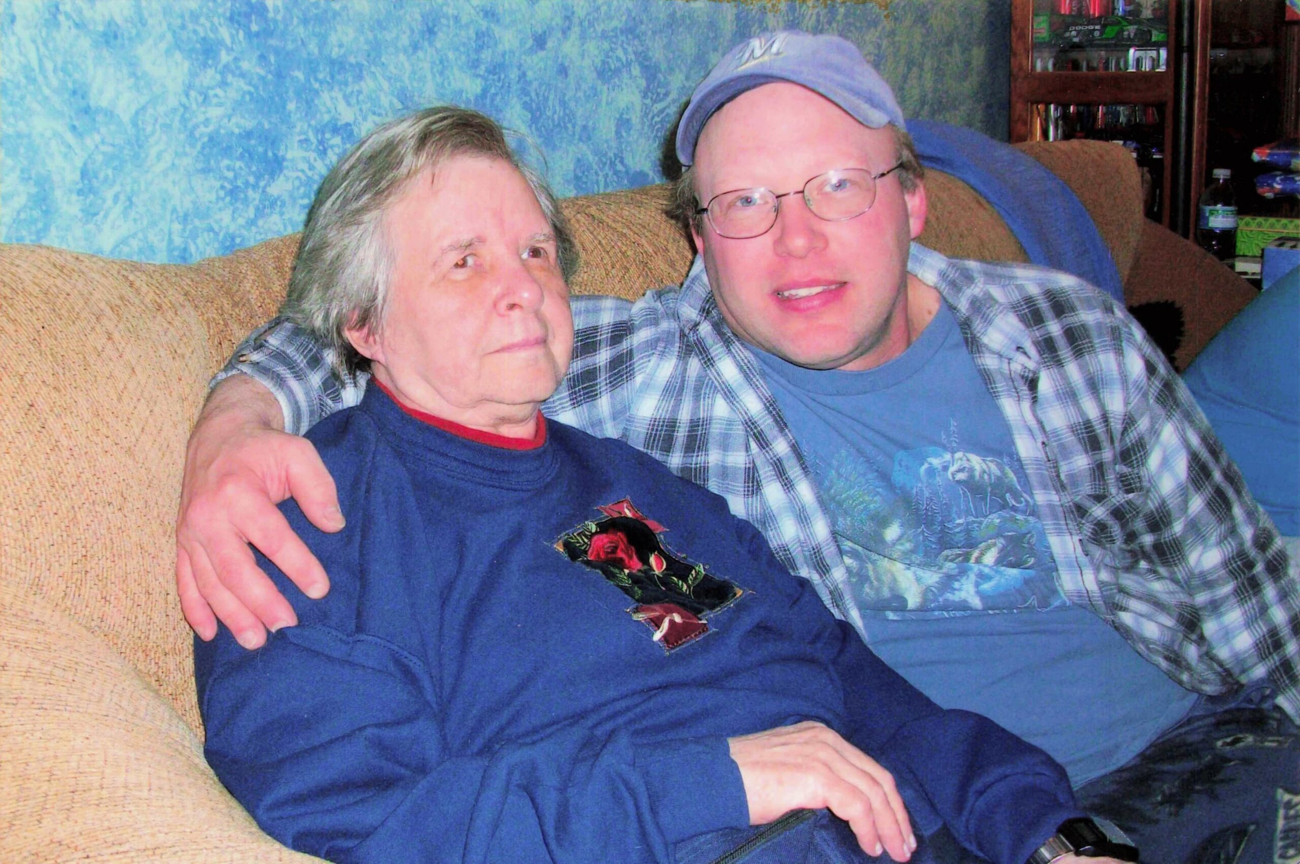 Two men sit tightly on a beige couch with one arm around the other, smiling at the camera in a living room setting.