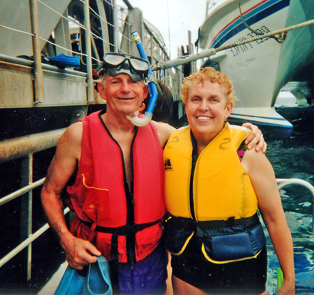Couple wearing life jackets and snorkeling gear on a dock near a boat, arm in arm and smiling at the camera.