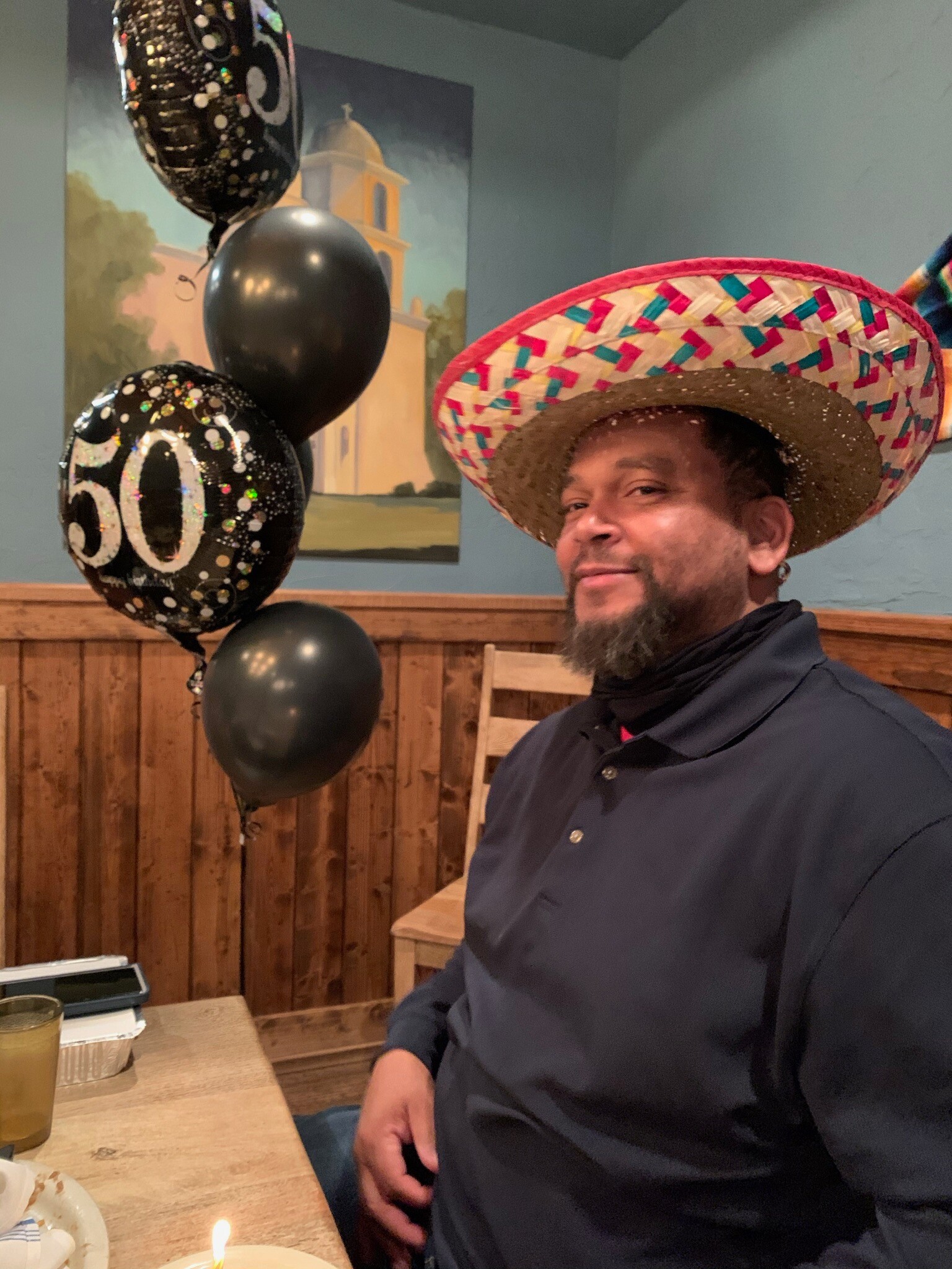 Man in a dark shirt wearing a colorful sombrero sits at a table with a lit candle and party balloons in the background.