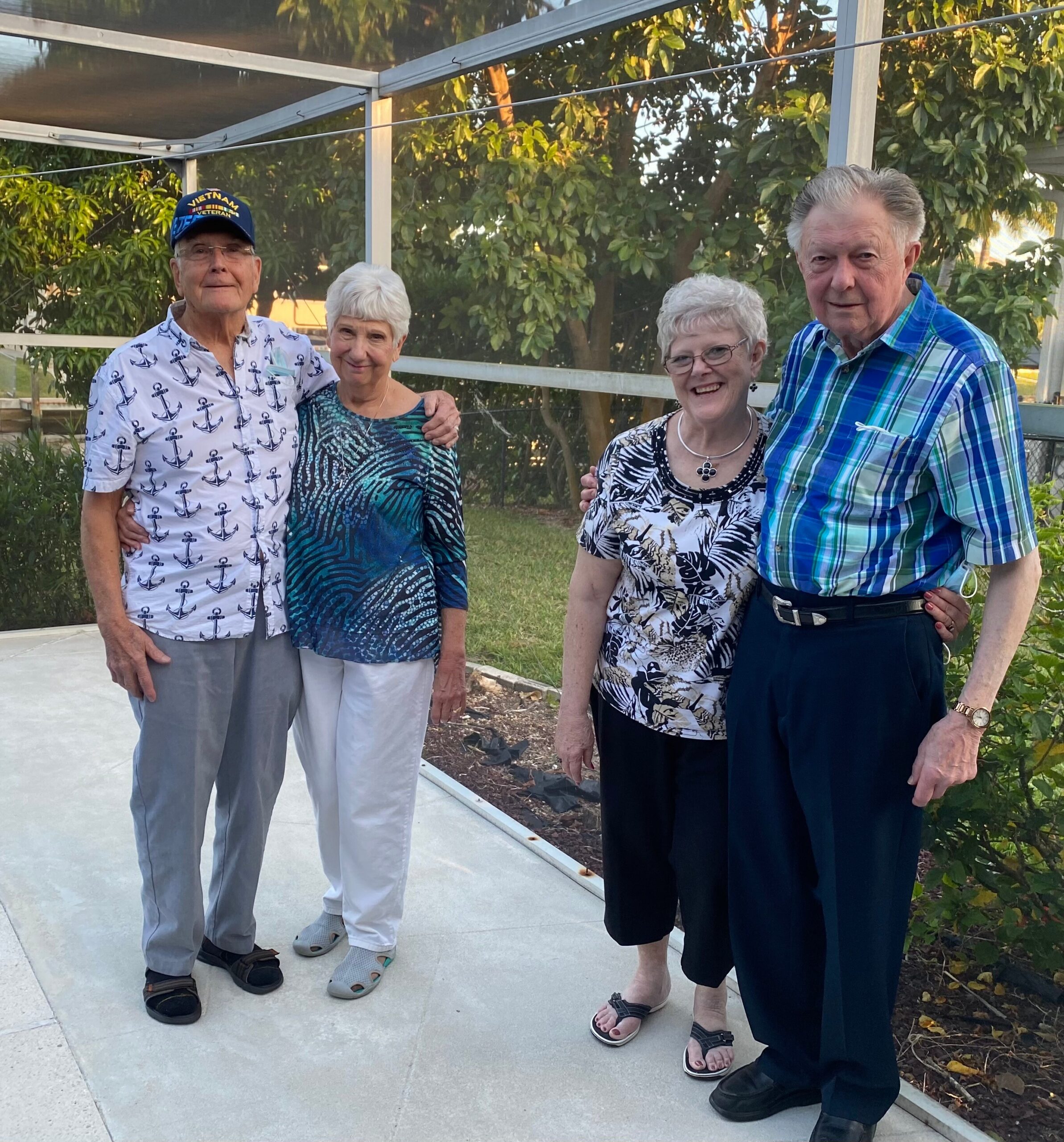 Four older adults pose together outdoors on a sunny patio with a glass railing and greenery in the background, smiling. 128 characters