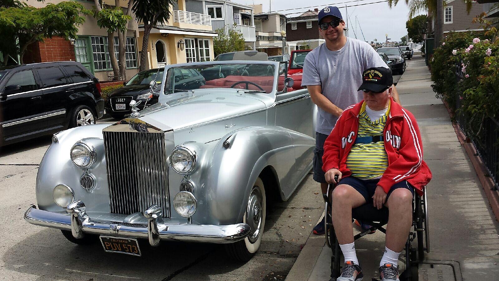 Silver vintage convertible parked on a residential street with two men, one in a wheelchair, posing beside it, smiling.