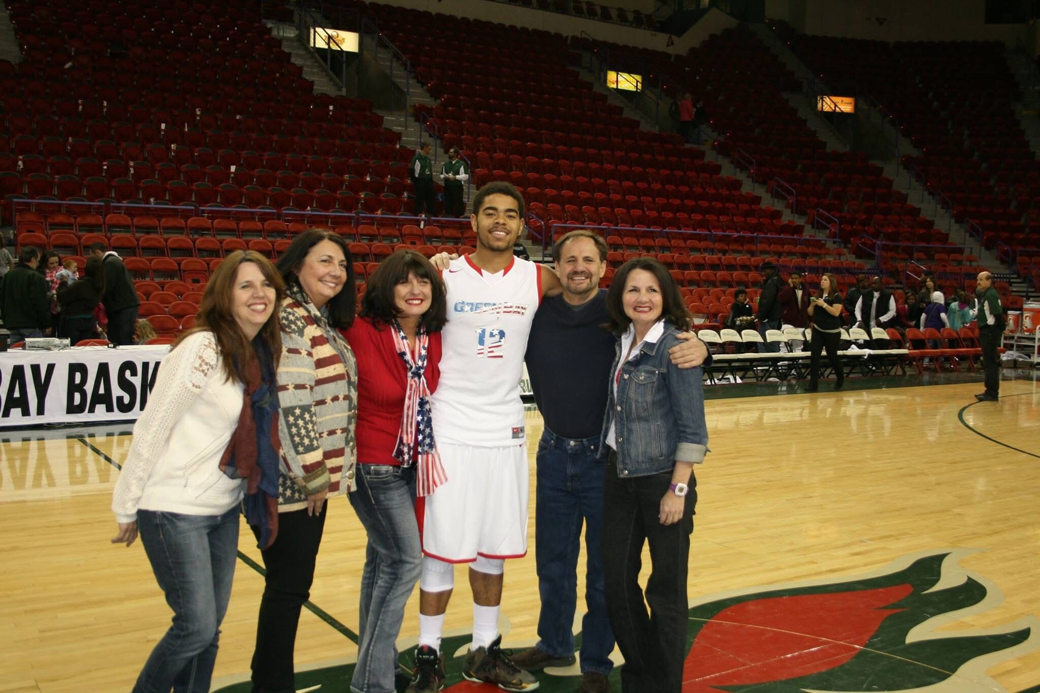 Six people pose together on a basketball court, including a player in a white uniform, with red arena seats behind them.