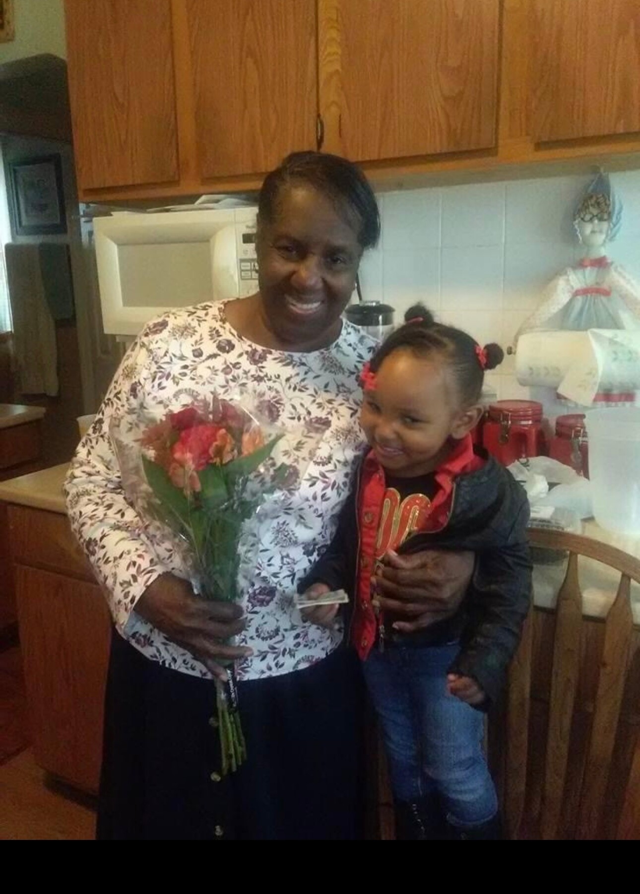 Smiling woman holding a bouquet of flowers with a young girl in a kitchen, posing together at home.