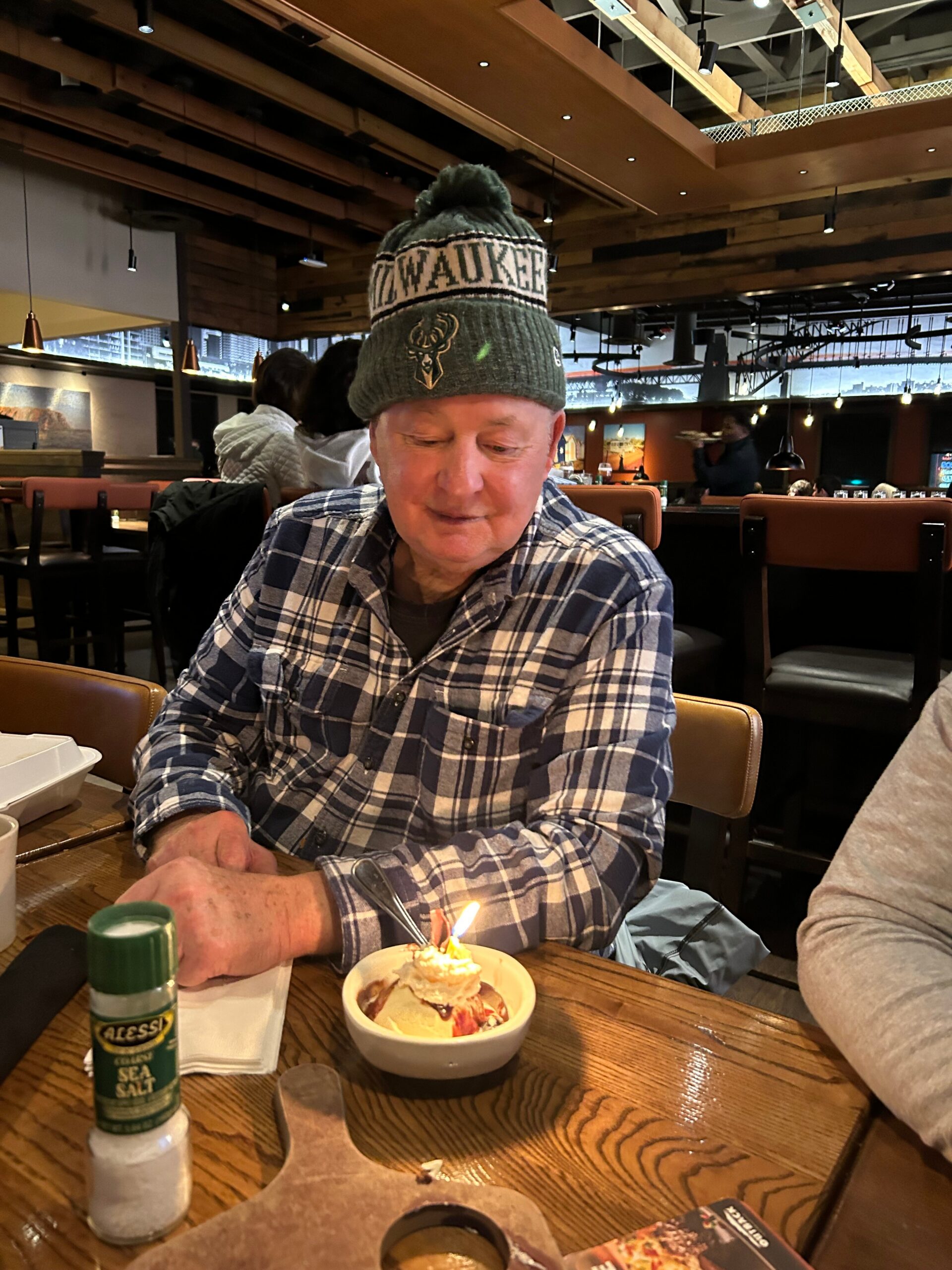 Older man in a plaid shirt wearing a Milwaukee Bucks beanie, looking at a small cake with a lit candle at a restaurant table.
