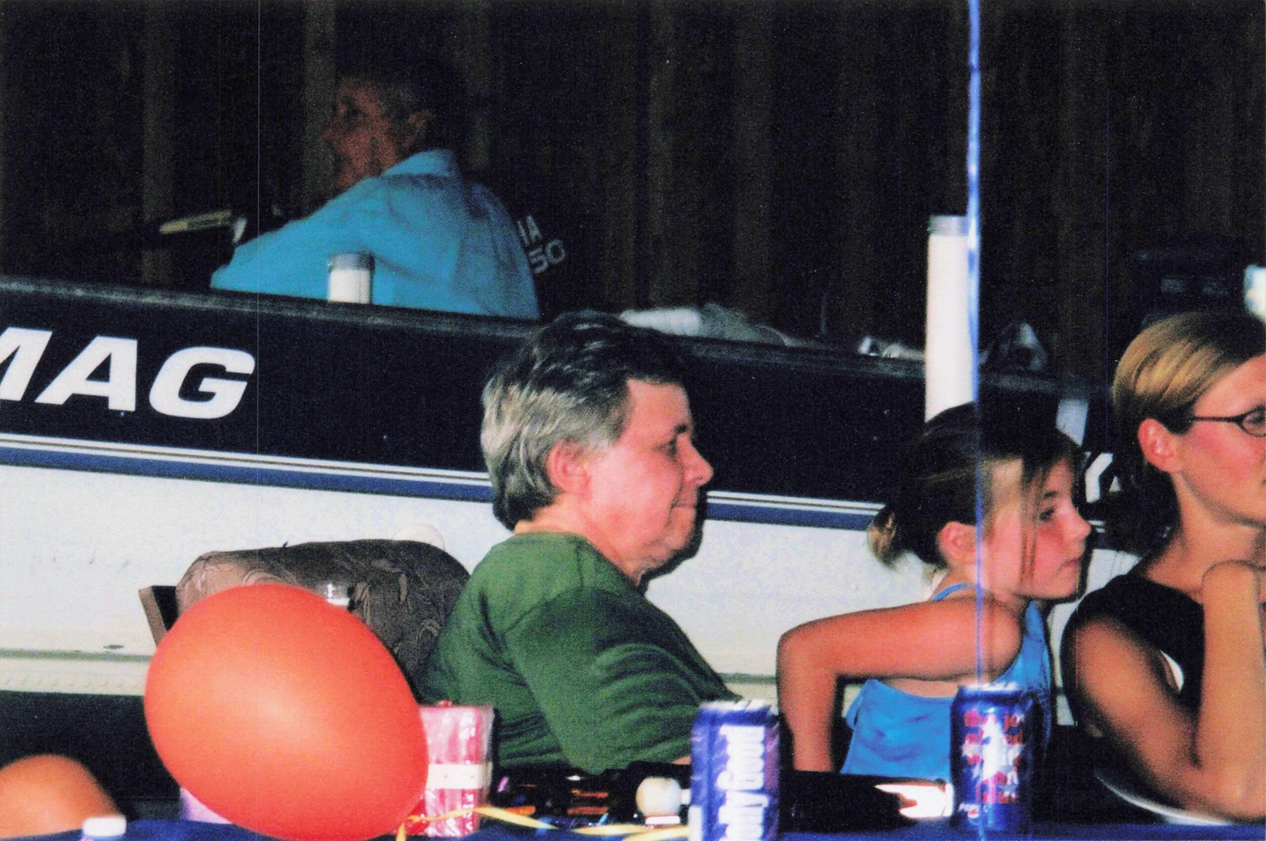 Group of people sitting around a table at a casual party, with soda cans and an orange balloon in the foreground.