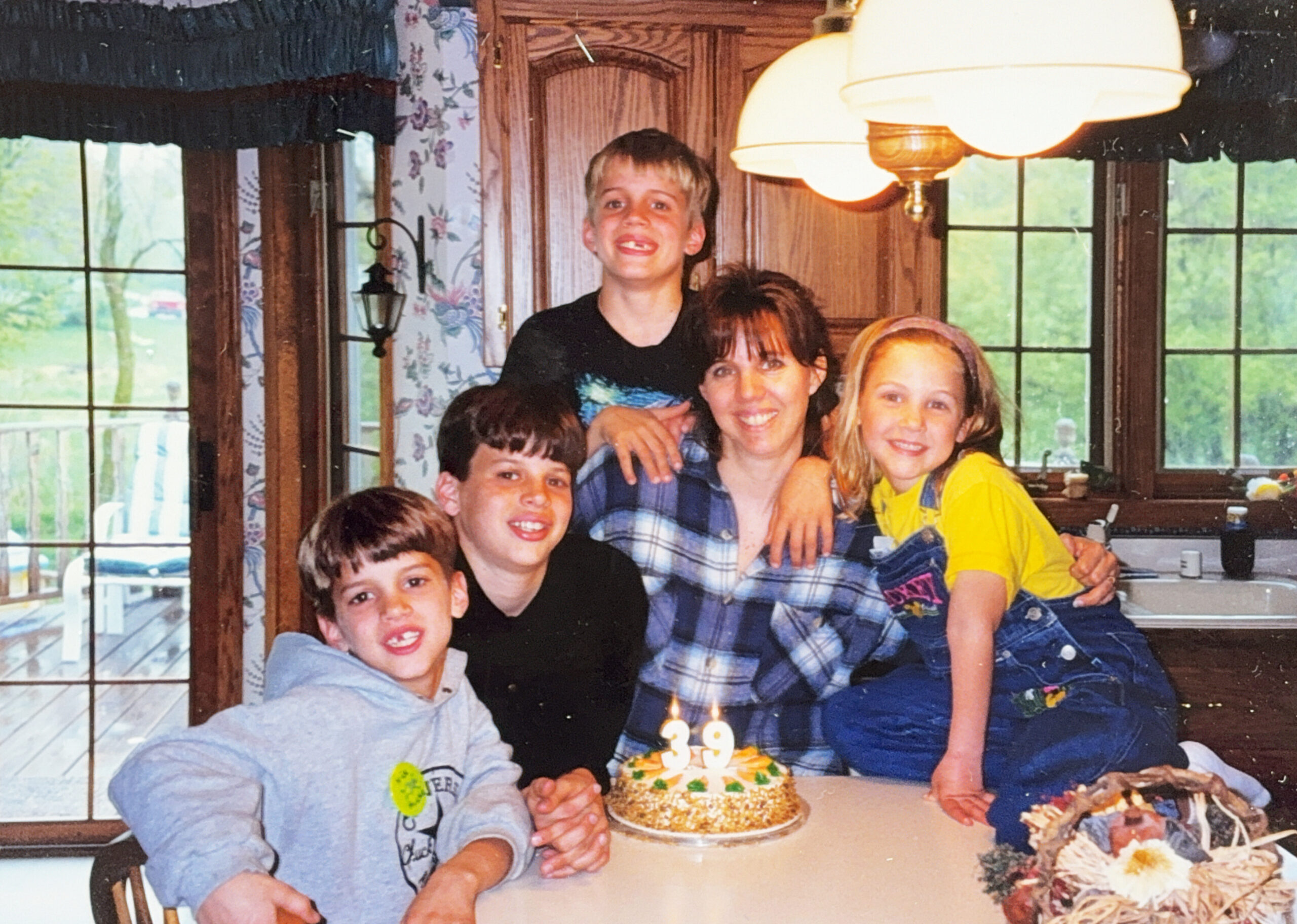 Family birthday celebration with kids and a woman gathered around a cake with lit candles at a home table.