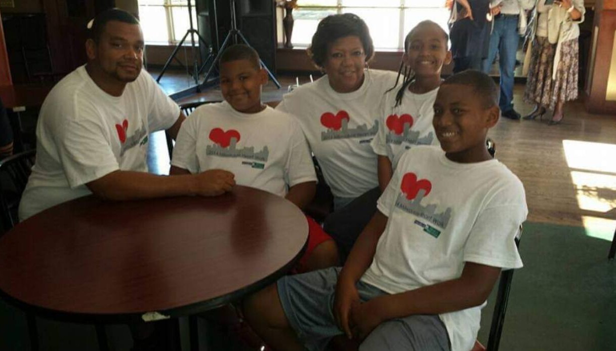 Five people sit around a round wooden table indoors, wearing white shirts with a red heart graphic and smiling at the camera.