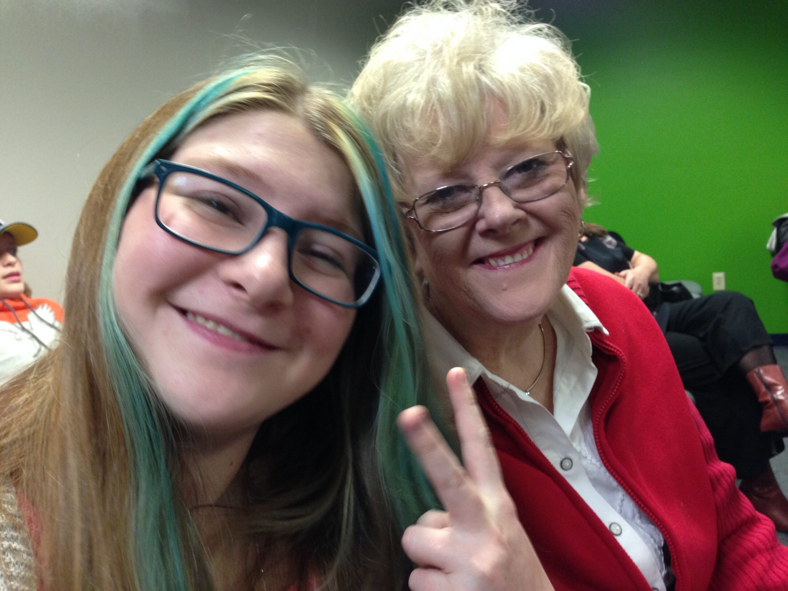 Close-up selfie of two smiling women: a younger woman with teal-highlighted hair and glasses on the left, and an older woman with short white hair and glasses on the right, in front of a green wall.