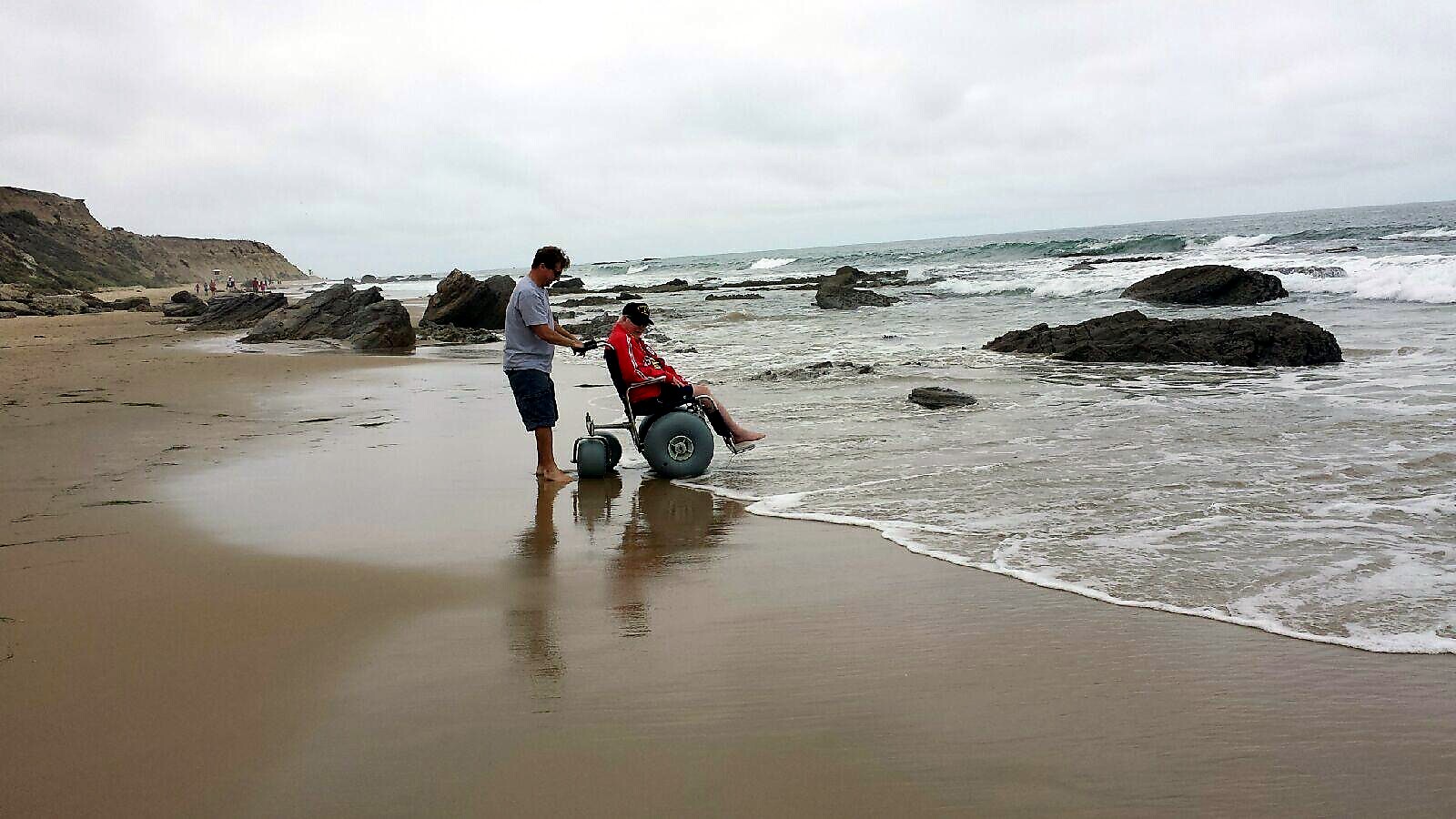Man assists a person in a red jacket seated in a beach wheelchair near the shoreline with waves and rocks in the background.