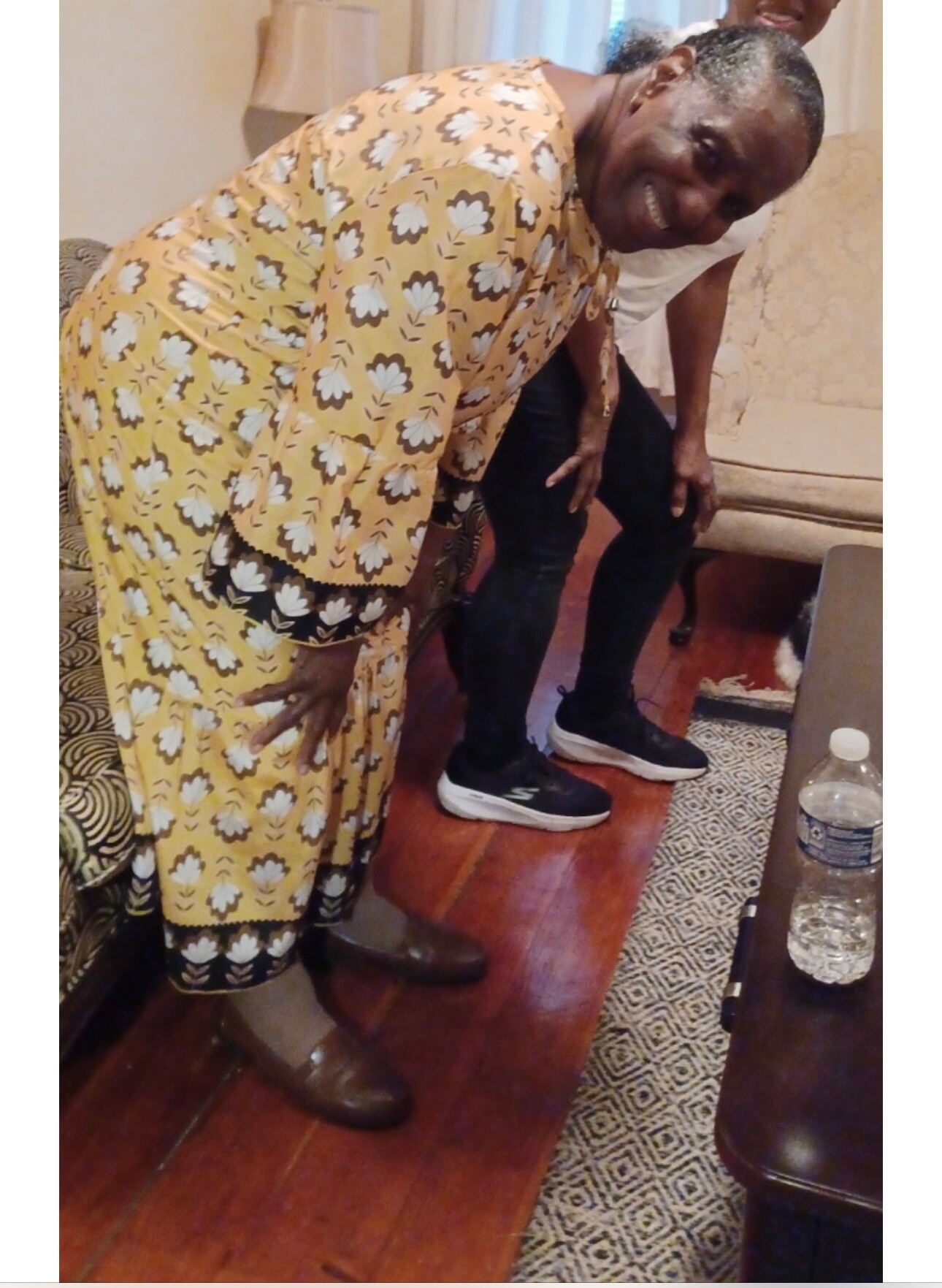 Woman in a yellow floral dress bends forward smiling, with a man standing behind her indoors; wooden floor and sofa visible, water bottle on a nearby table.