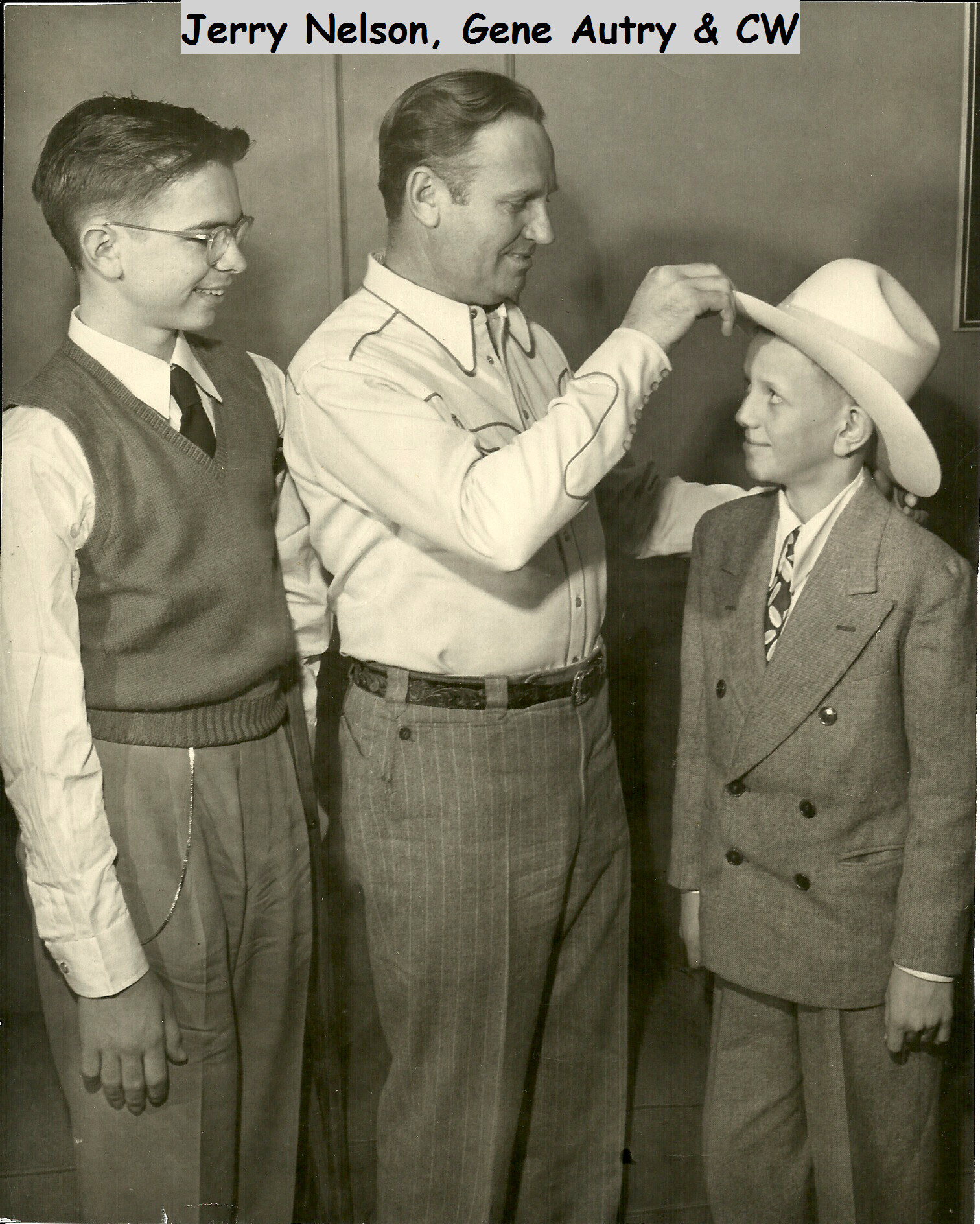 Three men in vintage clothing: a young man in a vest and tie on the left, a taller man in a western shirt in the middle adjusting a wide-brimmed cowboy hat onto a boy in a suit and tie, all looking at the hat with a smile.