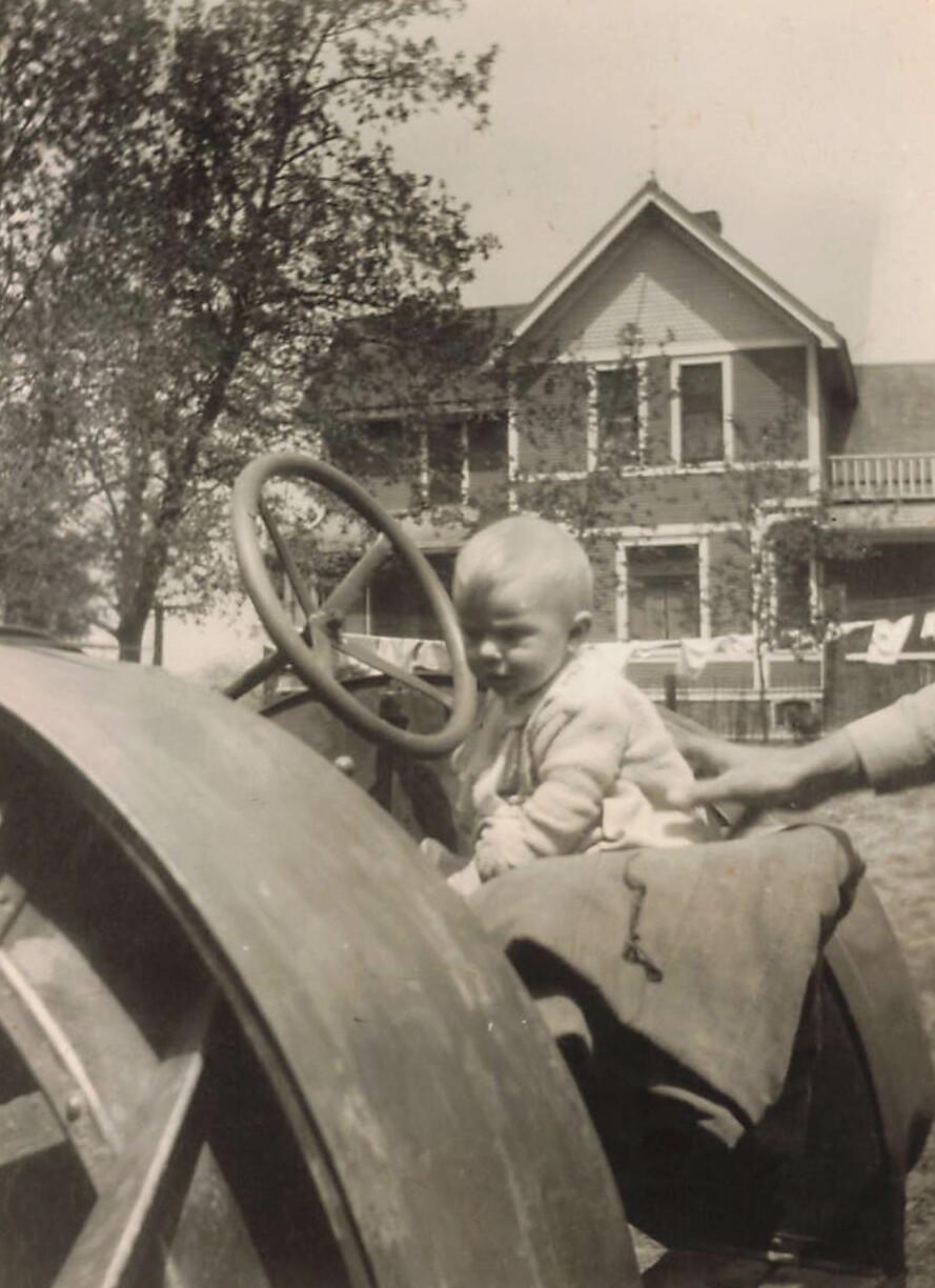 Baby sits on an old tractor steering wheel, supported by an adult's hand, with a house and trees in the background.