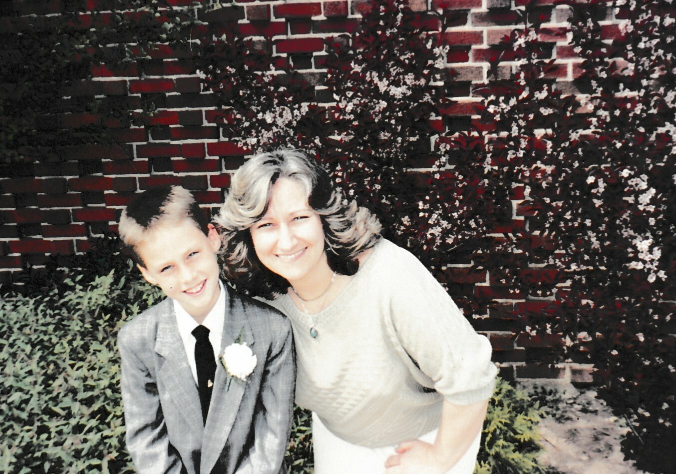 Woman and young boy smiling at the camera, posing outdoors in front of a brick wall and foliage, dressed nicely for a formal event.