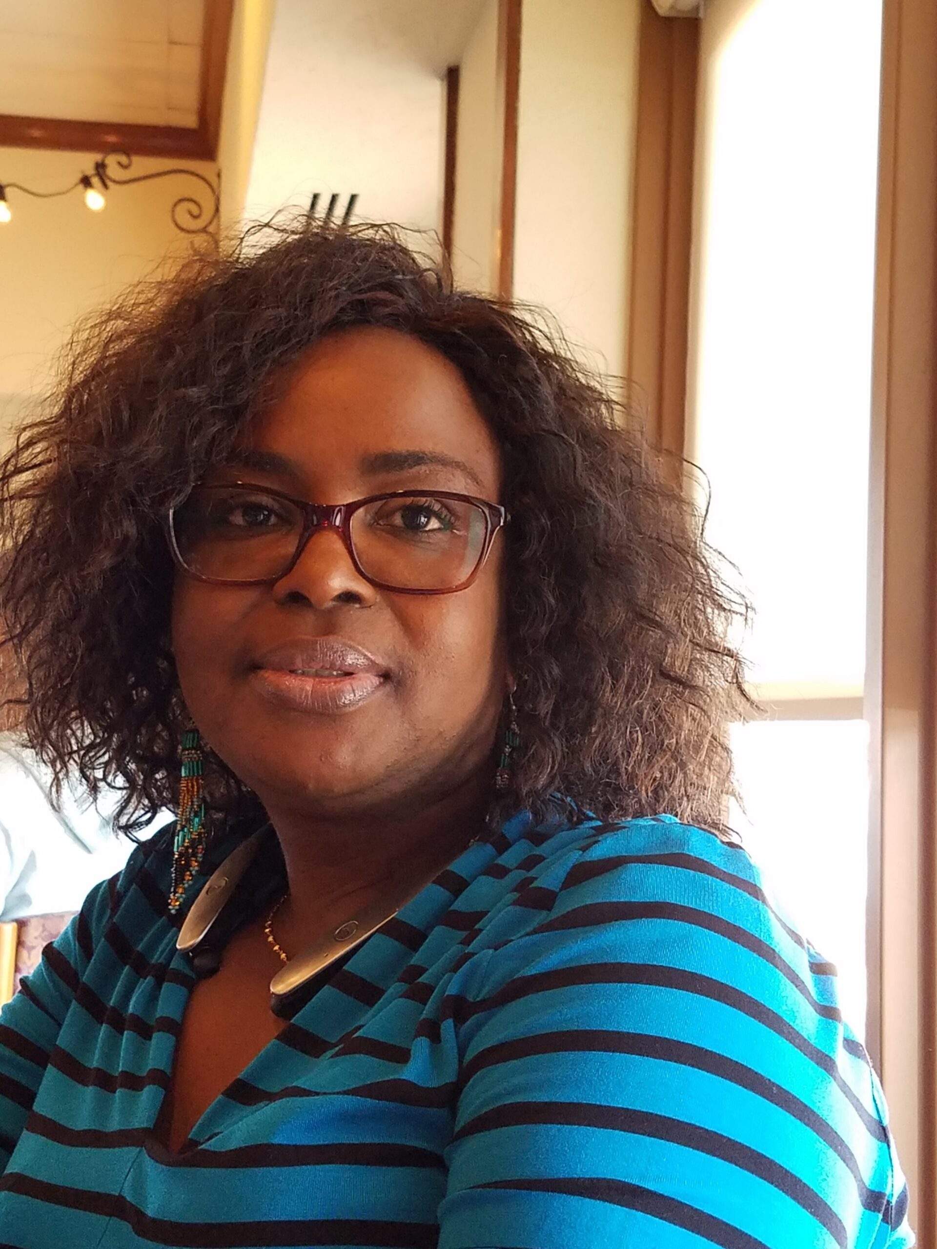 Woman with curly hair and glasses, wearing a turquoise and black striped top and beaded earrings, indoors.