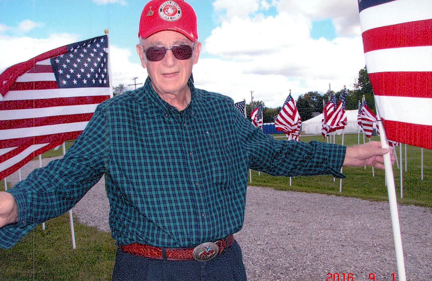 Older man wearing a red cap and sunglasses poses with arms outstretched among American flags at an outdoor patriotic display.