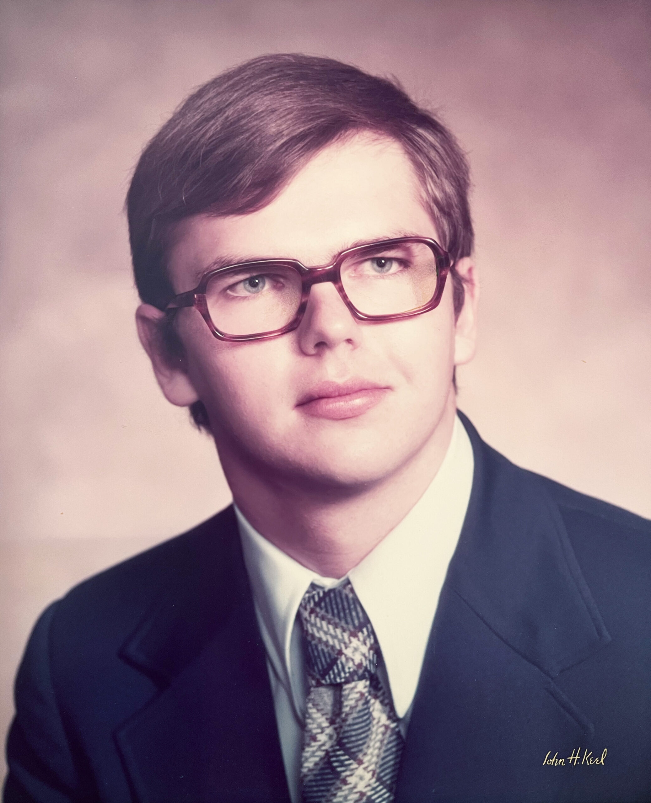 Head-and-shoulders portrait of a young man in a dark suit, white shirt, and patterned tie, wearing burgundy glasses.