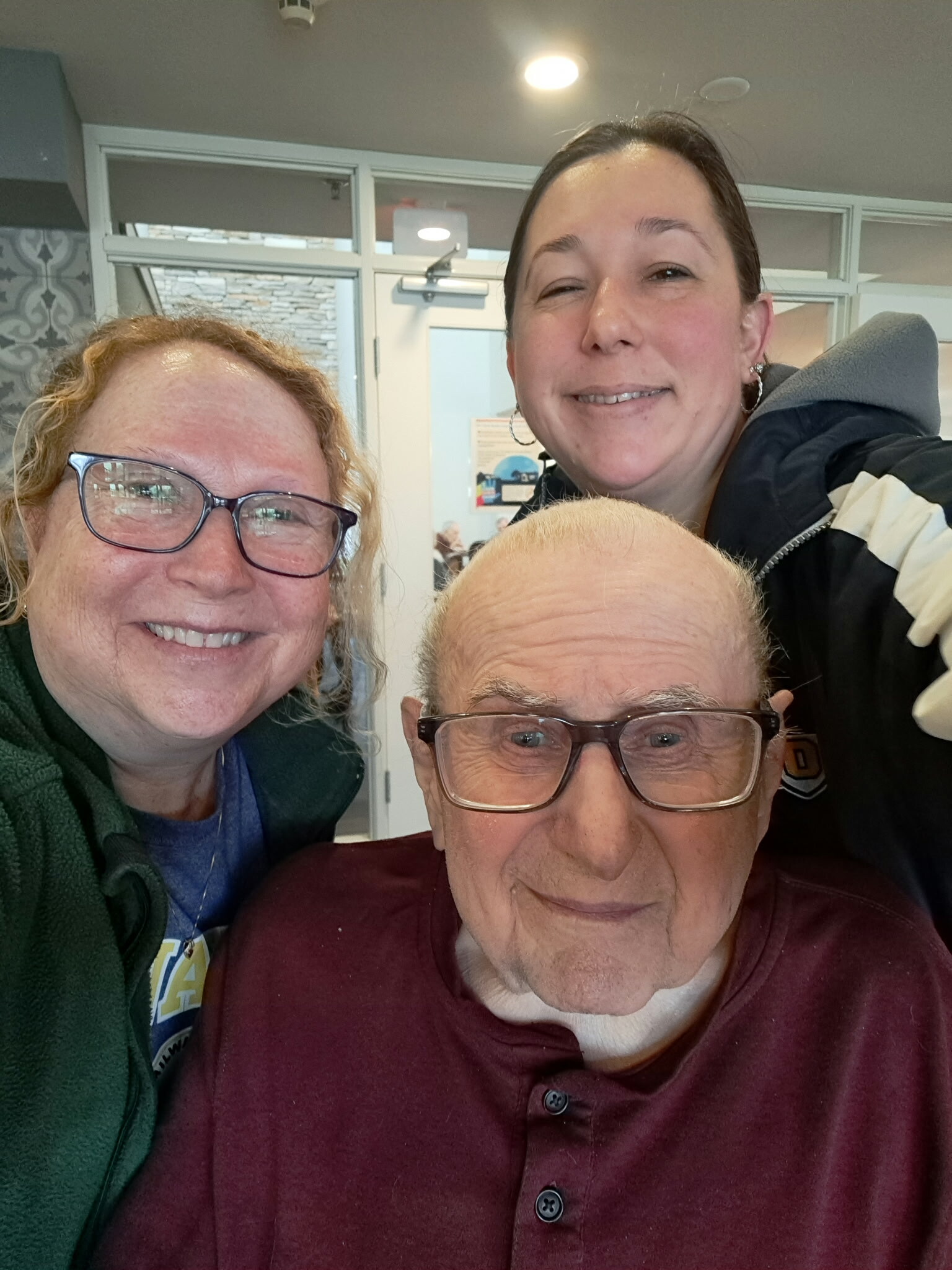 Smiling trio taking a selfie indoors; elderly man in front with glasses, two women behind him.