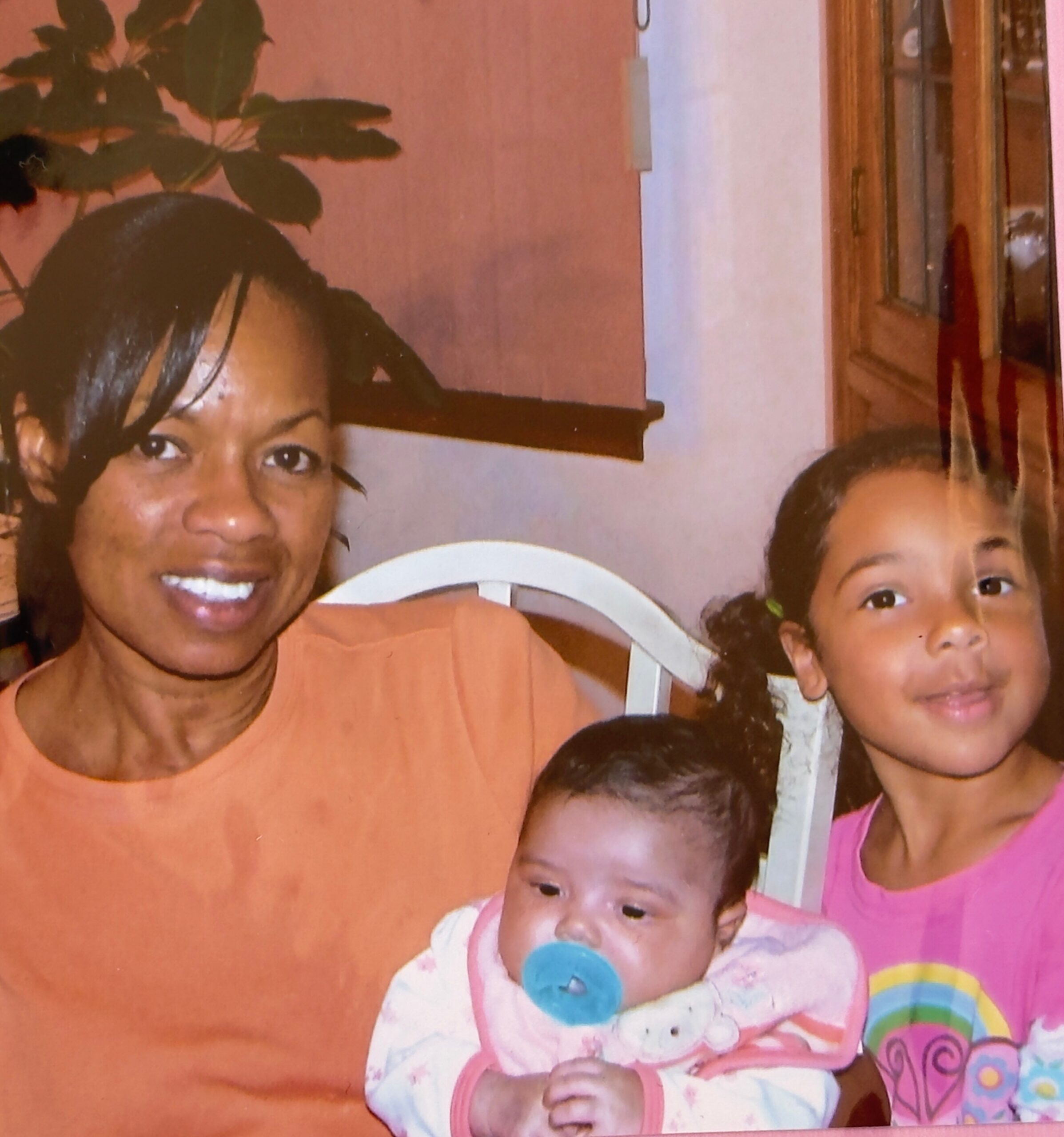 Close-up family photo of a woman with several young children in a warm living room, plant and cabinet in the background.