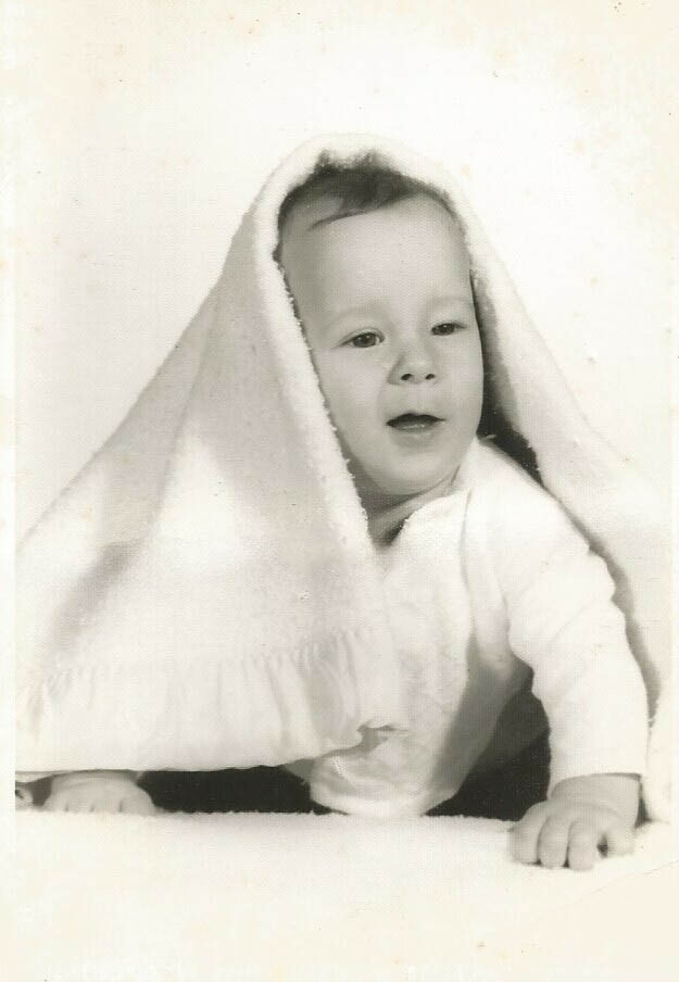 Infant with a white blanket draped over the head, peeking out and resting on hands and knees in a monochrome photo.