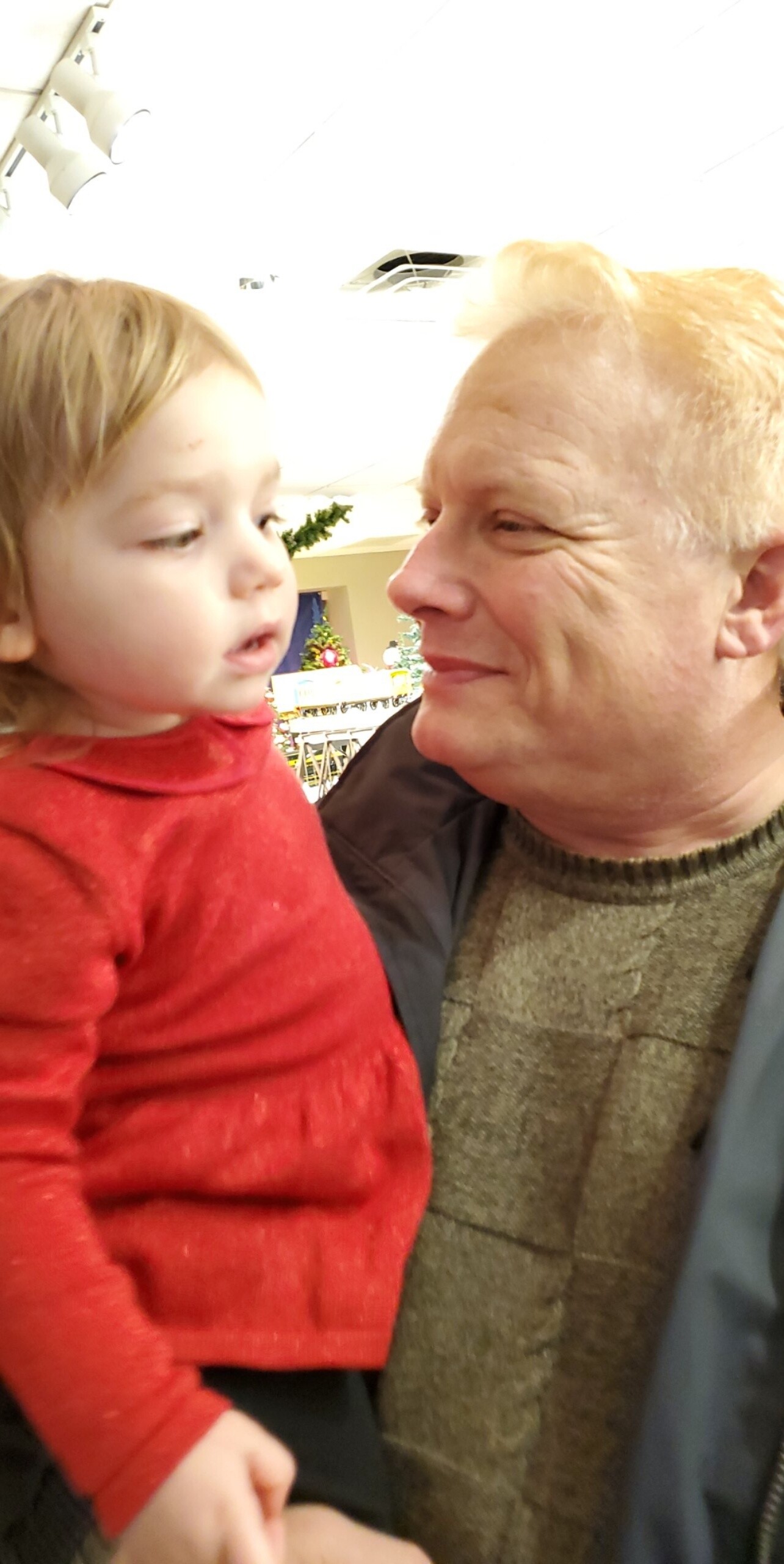 Grandfather smiling at a toddler he’s holding close indoors, with holiday decorations in the background.