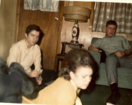 Three adults sit in a wood-paneled living room: two men on a sofa and a woman in the foreground (blurry), relaxed atmosphere.