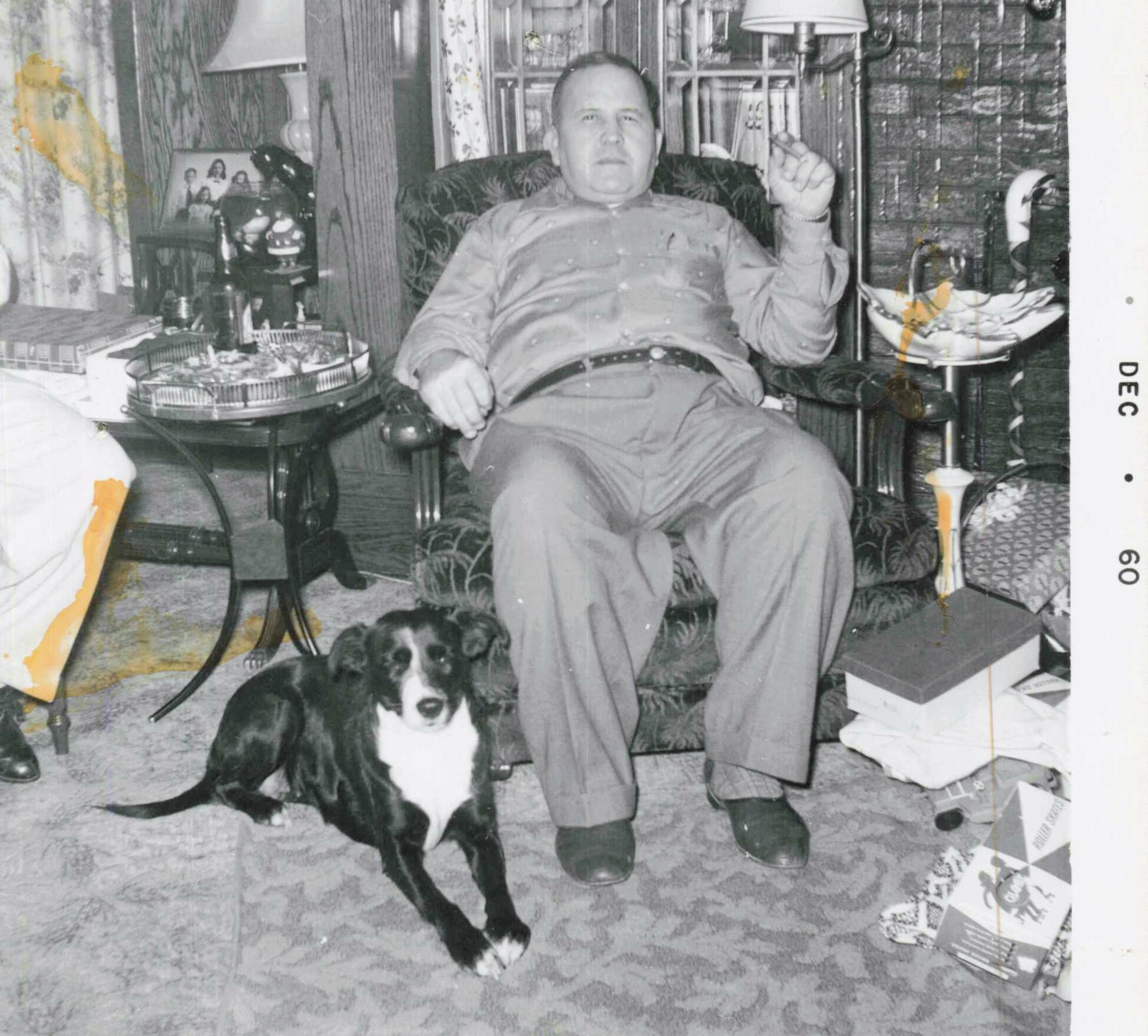 Older man sits in a patterned armchair with a black-and-white dog at his feet in a cluttered living room bedazzled with various items around him.