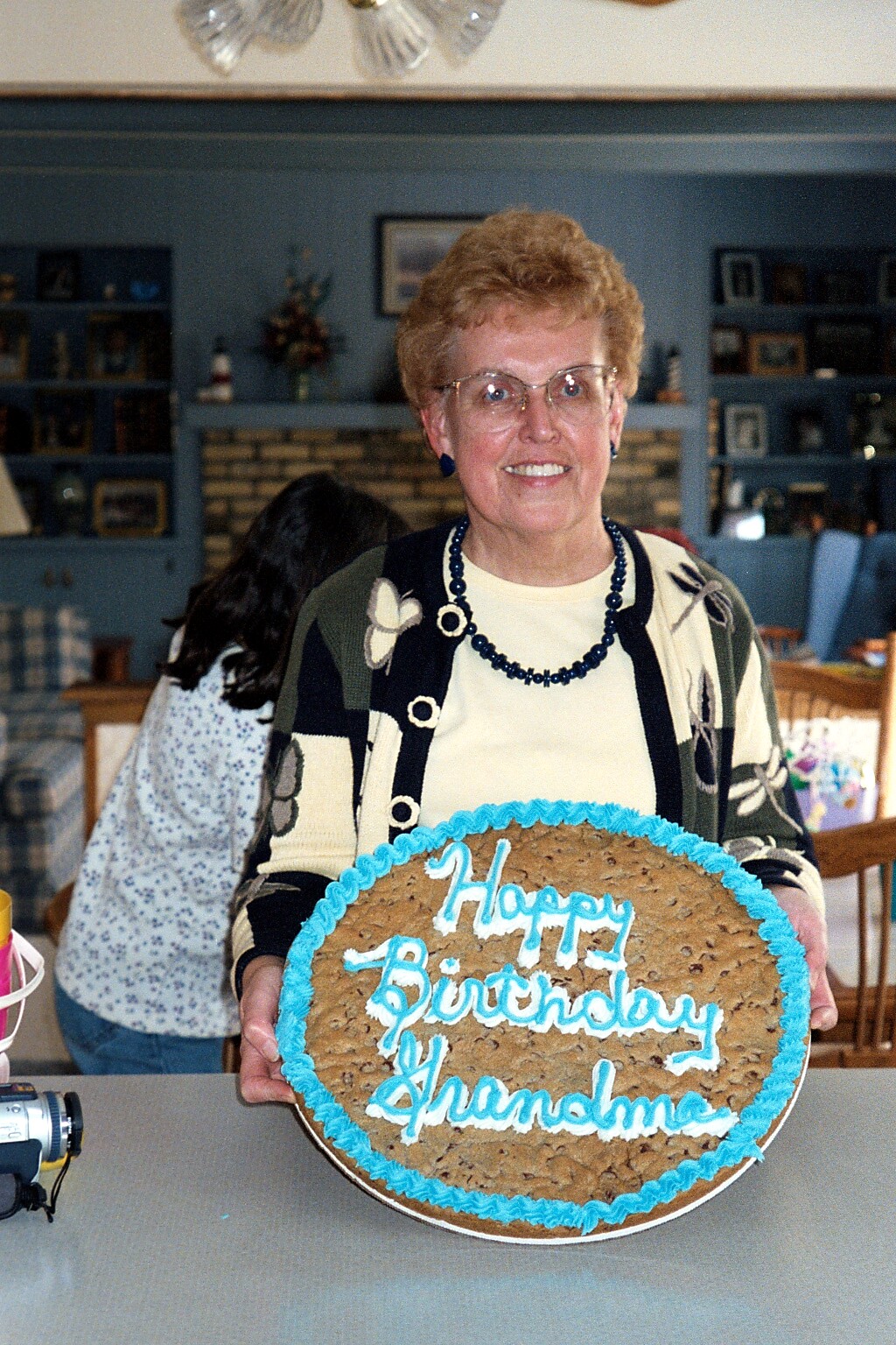 Older woman with glasses holds a round birthday cake with blue icing and the words 'Happy Birthday Grandmom' on it.