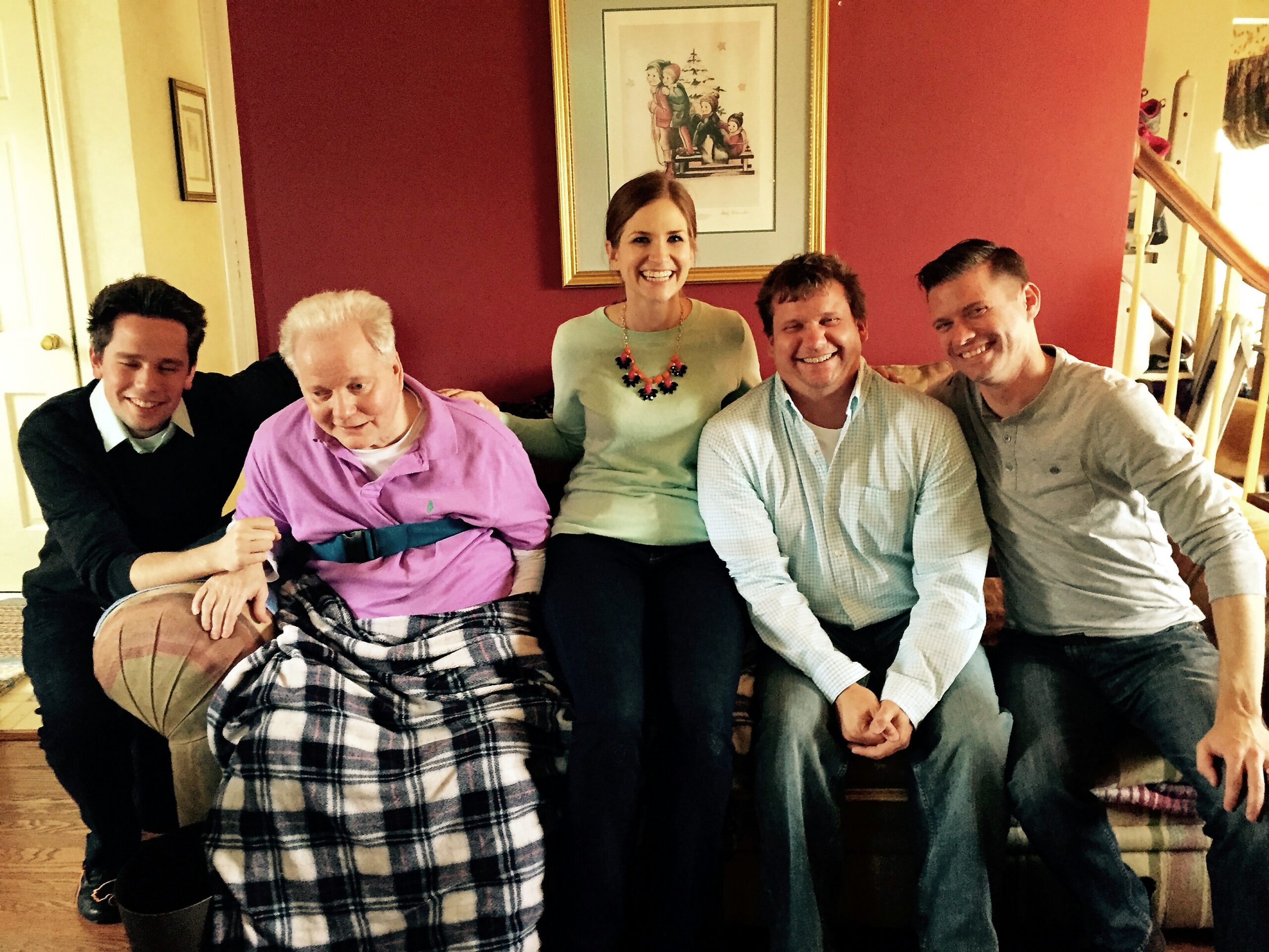 Five people sitting on a sofa in a cozy living room, smiling at the camera with a plaid blanket on the lap. A framed picture hangs on a red wall behind them.