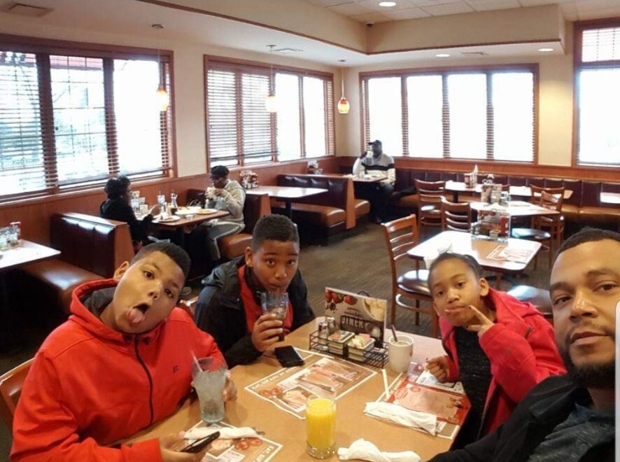 A man and three kids pose for a selfie at a casual restaurant table with drinks, cups, and menus visible.